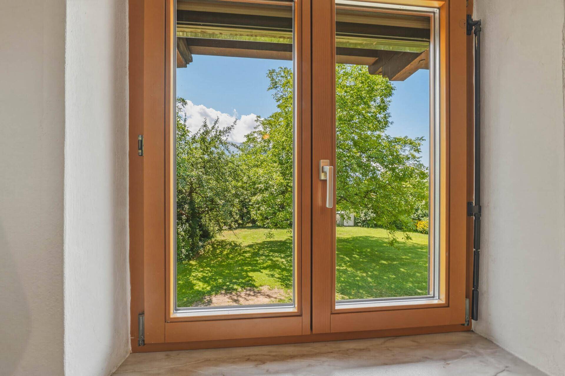 Fenster mit Blick auf den grünen Garten und die umliegende Natur an einem sonnigen Tag.