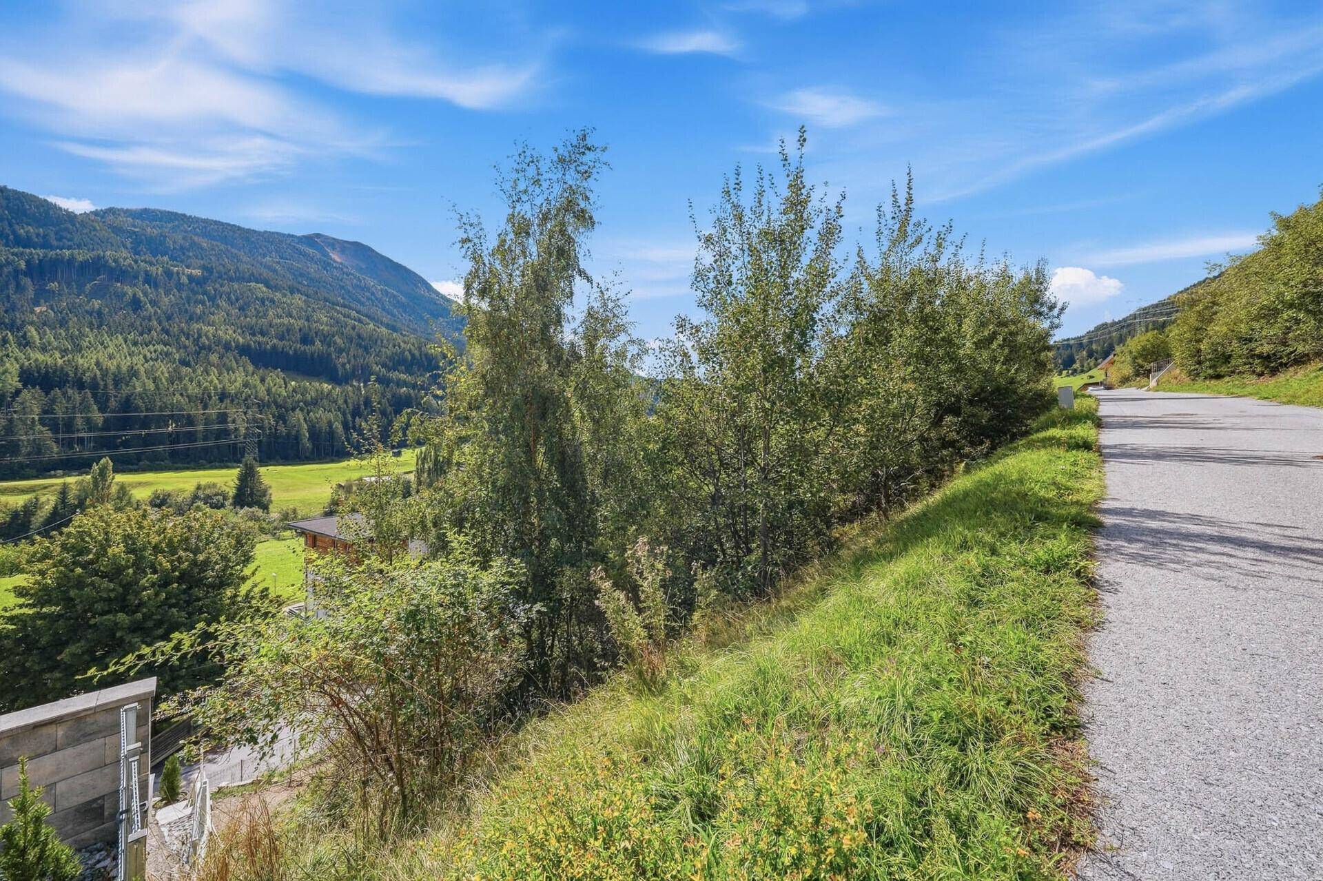 Blick auf eine Landstraße, umgeben von grünen Hängen und Bäumen, mit einem weiten Bergtal im Hintergrund.