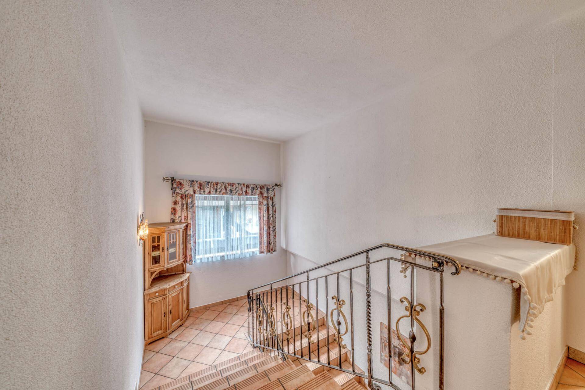 Treppenhaus mit schmiedeeisernem Geländer, Terrakottafliesen und einem kleinen Fenster mit Vorhängen.