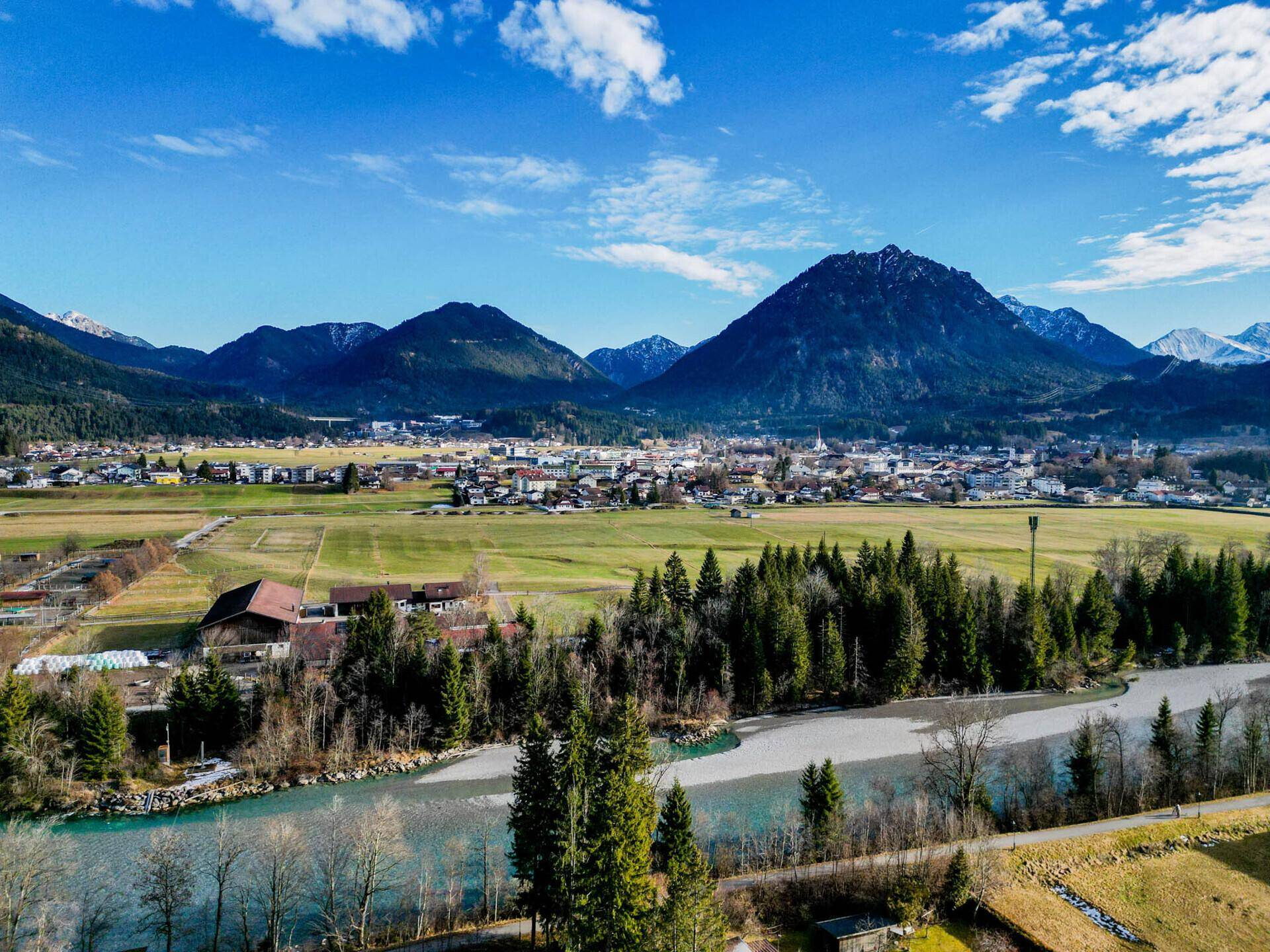 Weitläufige Landschaftsaufnahme mit einem Fluss, einer Stadt in der Ferne und einer beeindruckenden Bergkulisse.
