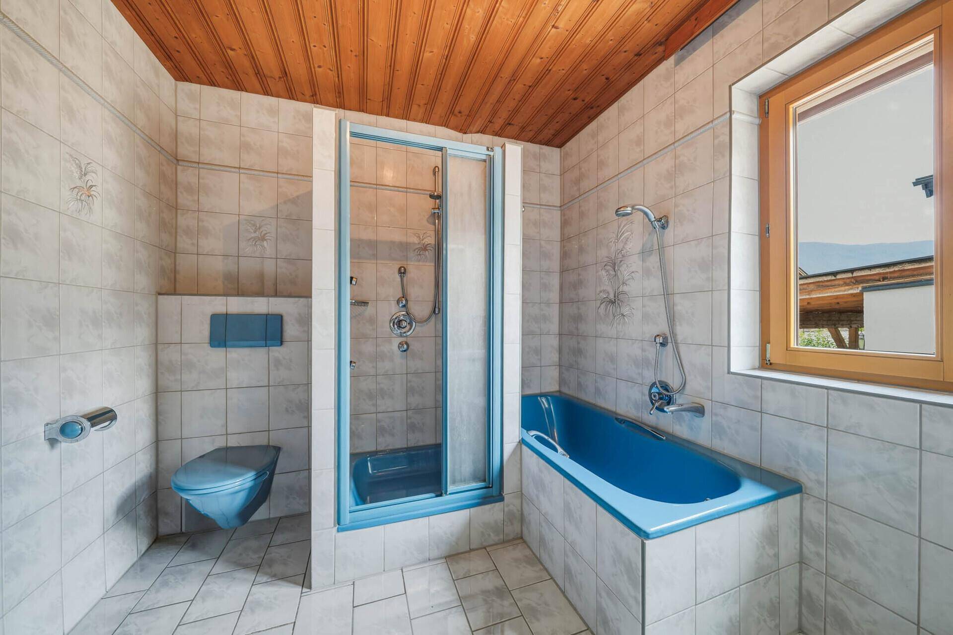 Badezimmer mit blauer Badewanne, Dusche, WC und gefliesten Wänden sowie einer Holzdecke.