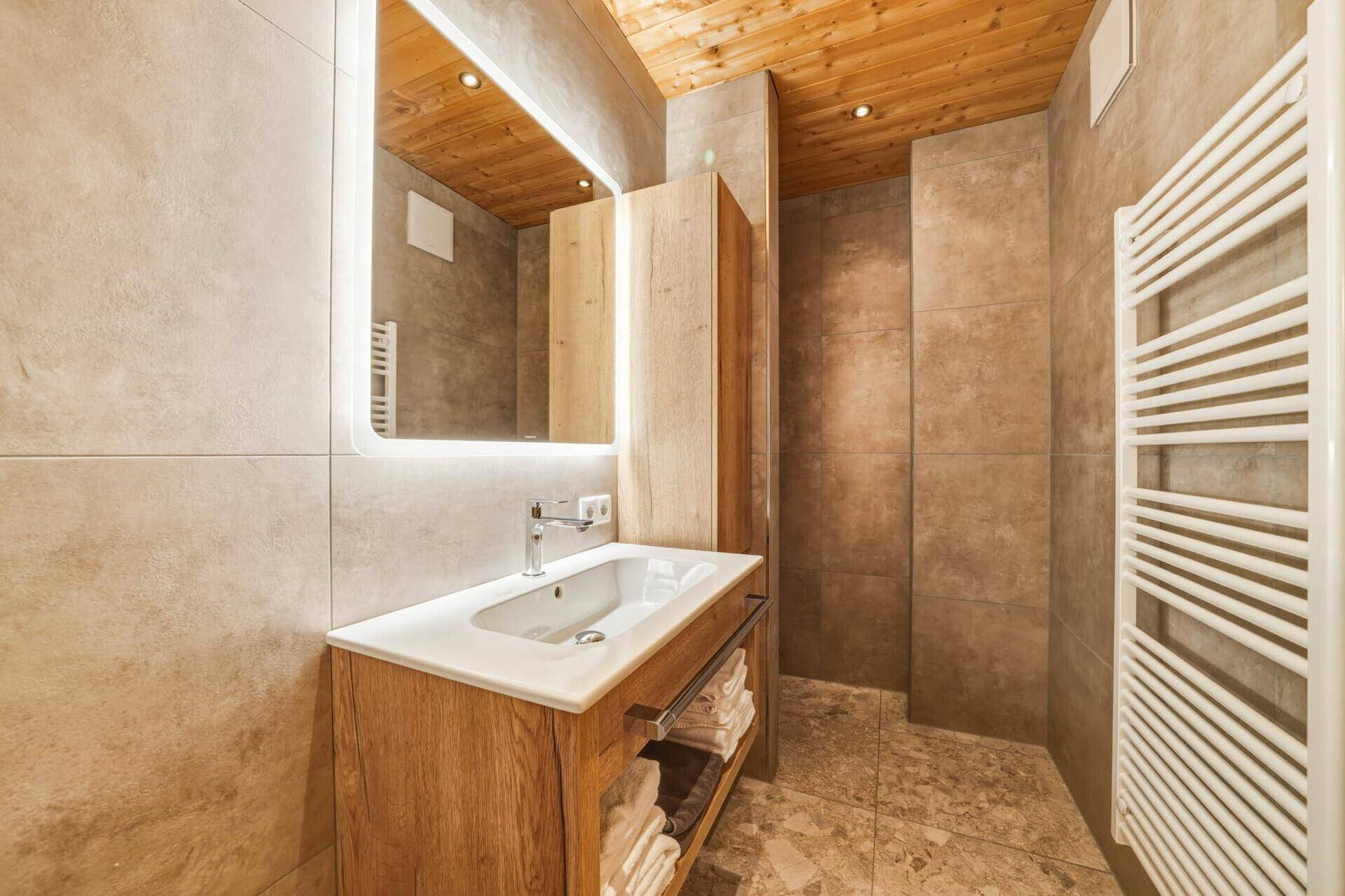 Modernes Badezimmer mit Holzbalkendecke, großformatigen Fliesen und beleuchtetem Spiegel.