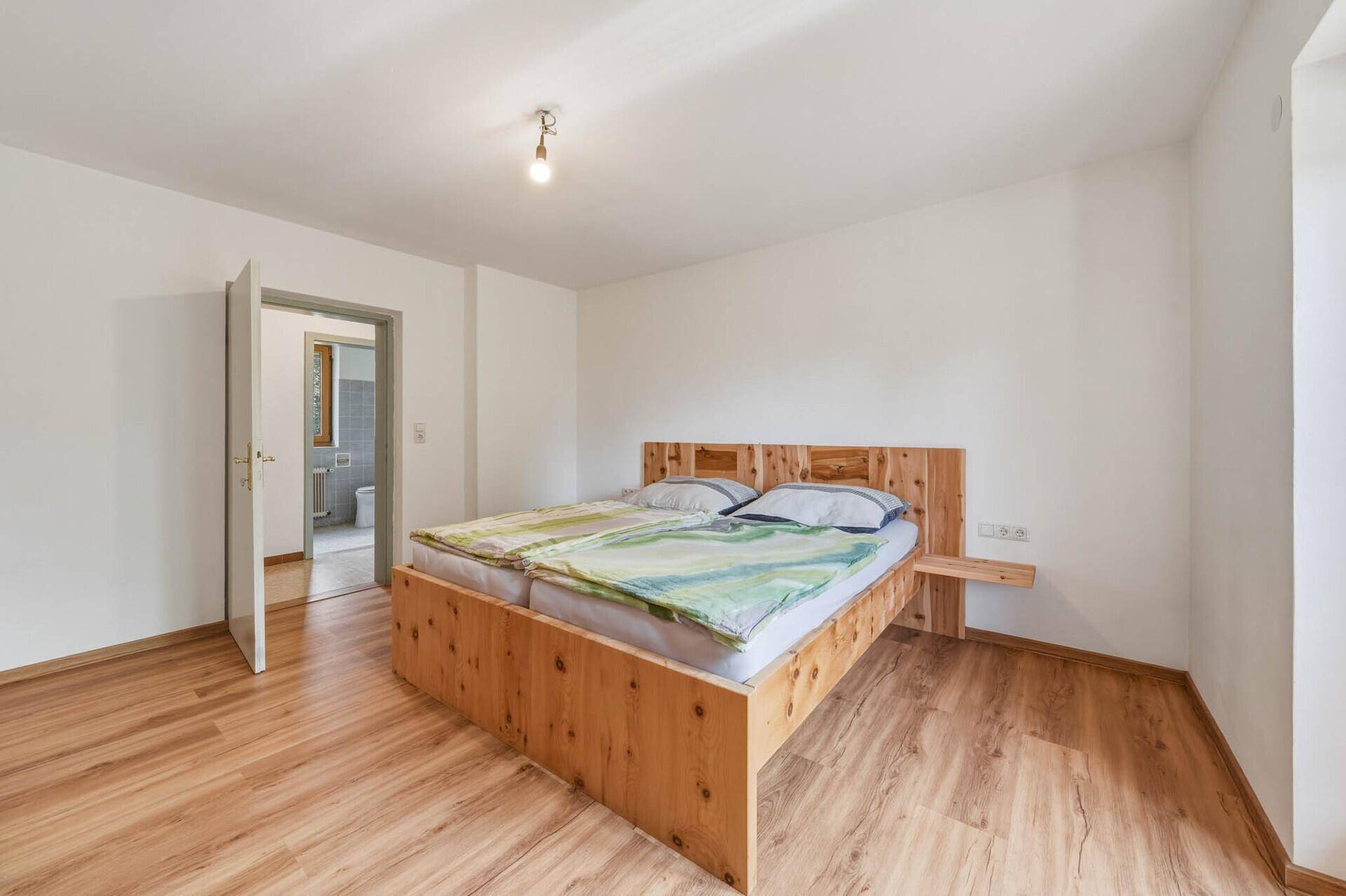 Schlafzimmer mit Holzbett, Laminatboden und weiß gestrichenen Wänden, mit Zugang zum Badezimmer.