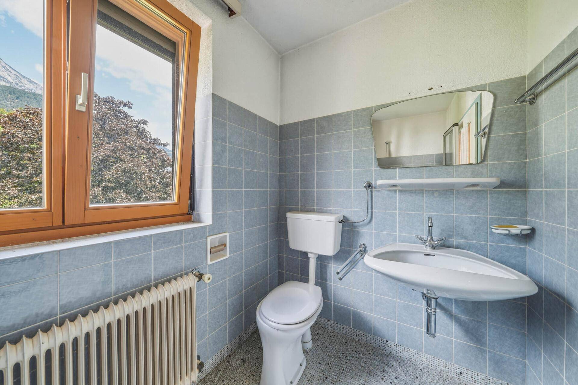 Badezimmer im 80er-Jahre-Stil mit blauen Fliesen, WC, Waschbecken und Fenster.
