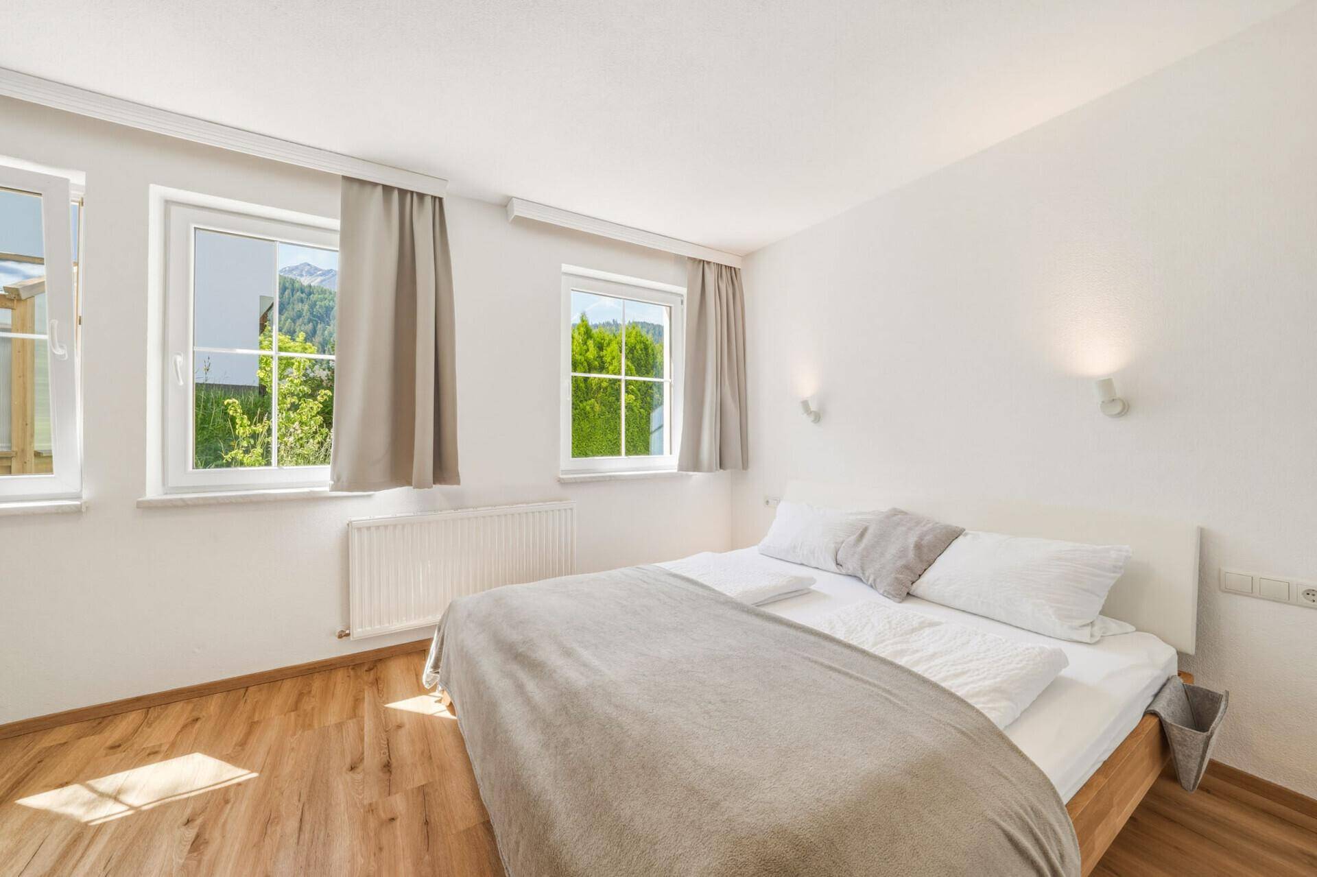 Helles Schlafzimmer mit Holzboden, Doppelbett und Fenstern mit Blick auf grüne Bäume und Berge.