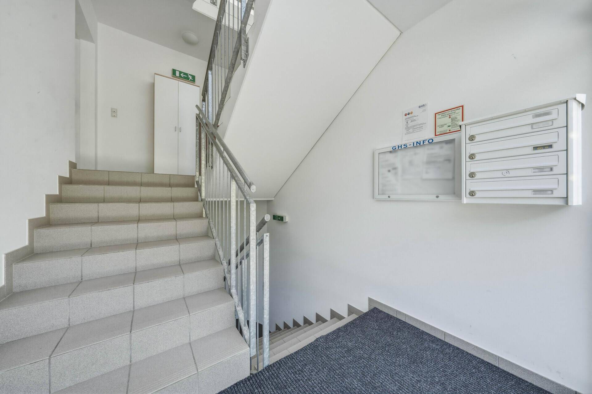 Helles Treppenhaus mit gefliesten Stufen, Metallgeländer und Briefkästen an der Wand.