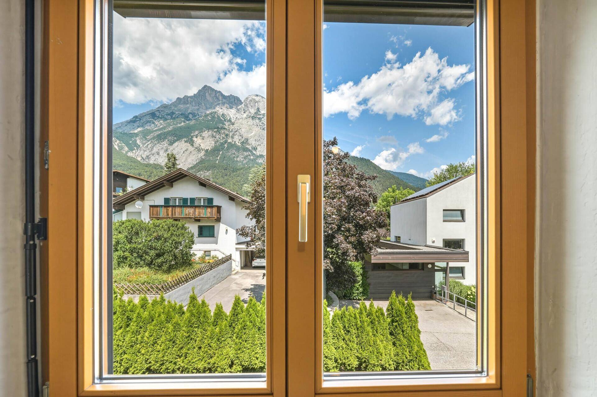 Blick aus einem Fenster auf benachbarte Häuser, gepflegte Hecken und eine beeindruckende Bergkulisse.