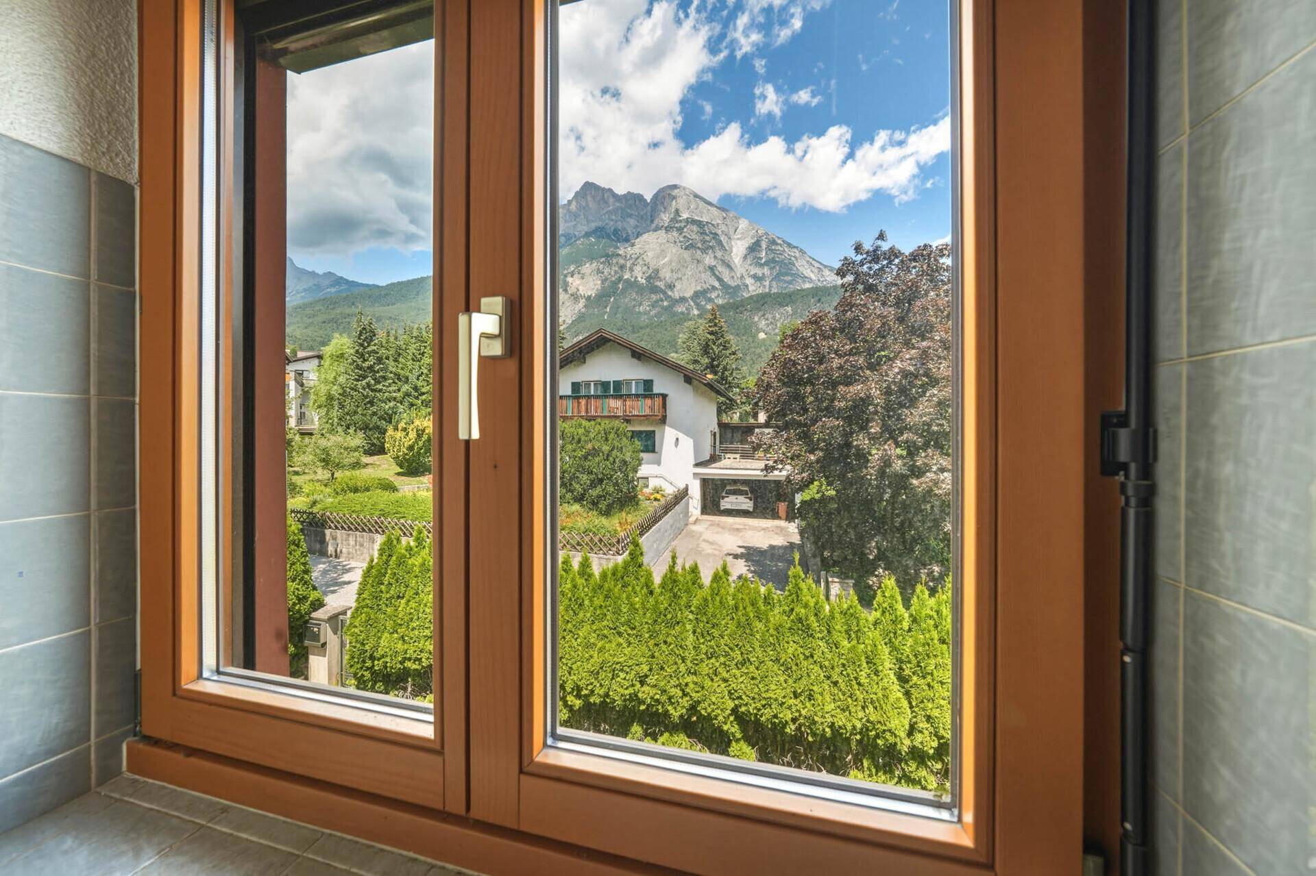 Blick aus einem Fenster auf benachbarte Häuser, grüne Hecken und eine beeindruckende Bergkulisse.