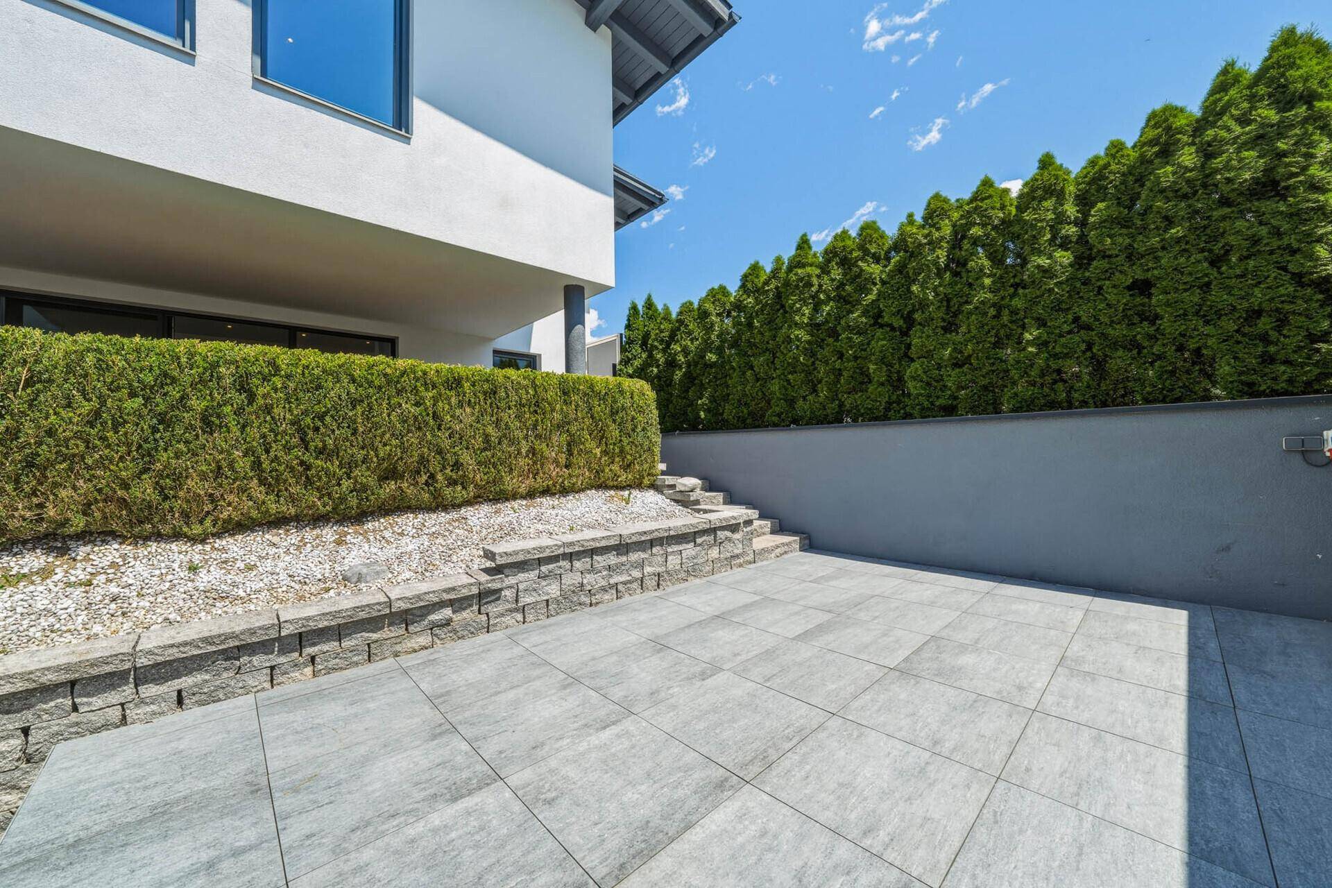 Gepflasterter Außenbereich mit Stützmauern, einer hohen Hecke und dem Haus im Hintergrund.