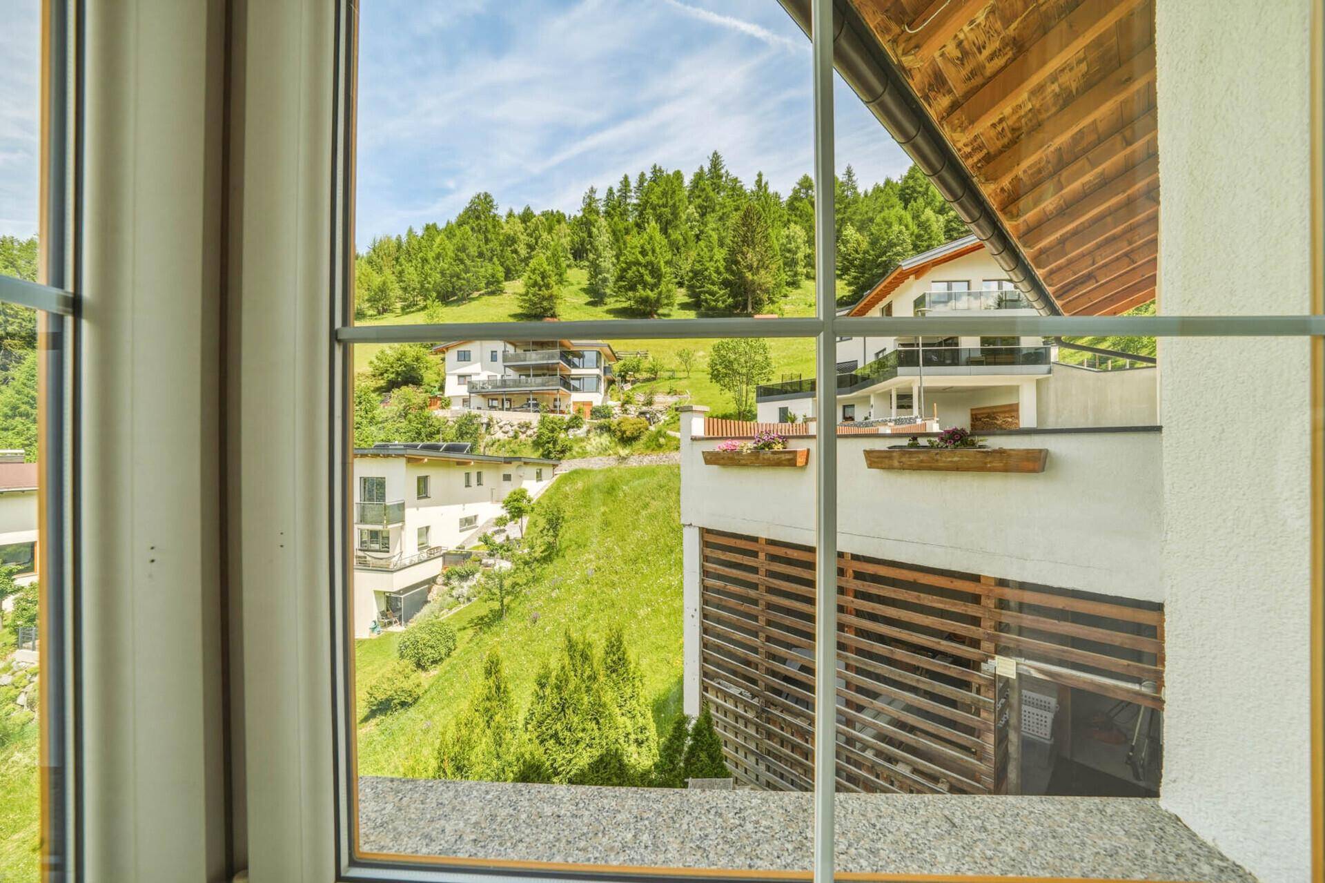 Ausblick aus dem Fenster auf Häuser am Hang, umgeben von grünen Bäumen und Bergen.