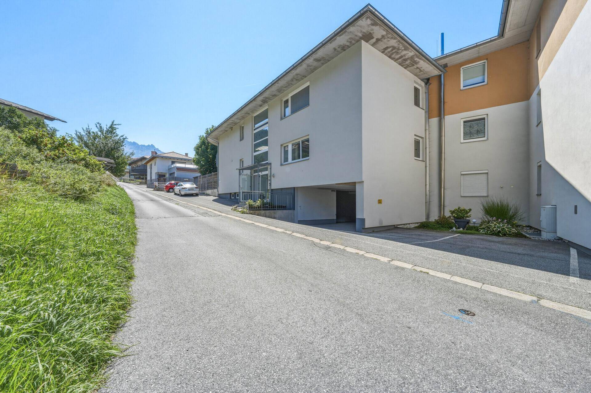 Gesamtansicht des modernen Gebäudes von der Straße, mit Tiefgaragenzufahrt und Bergblick.