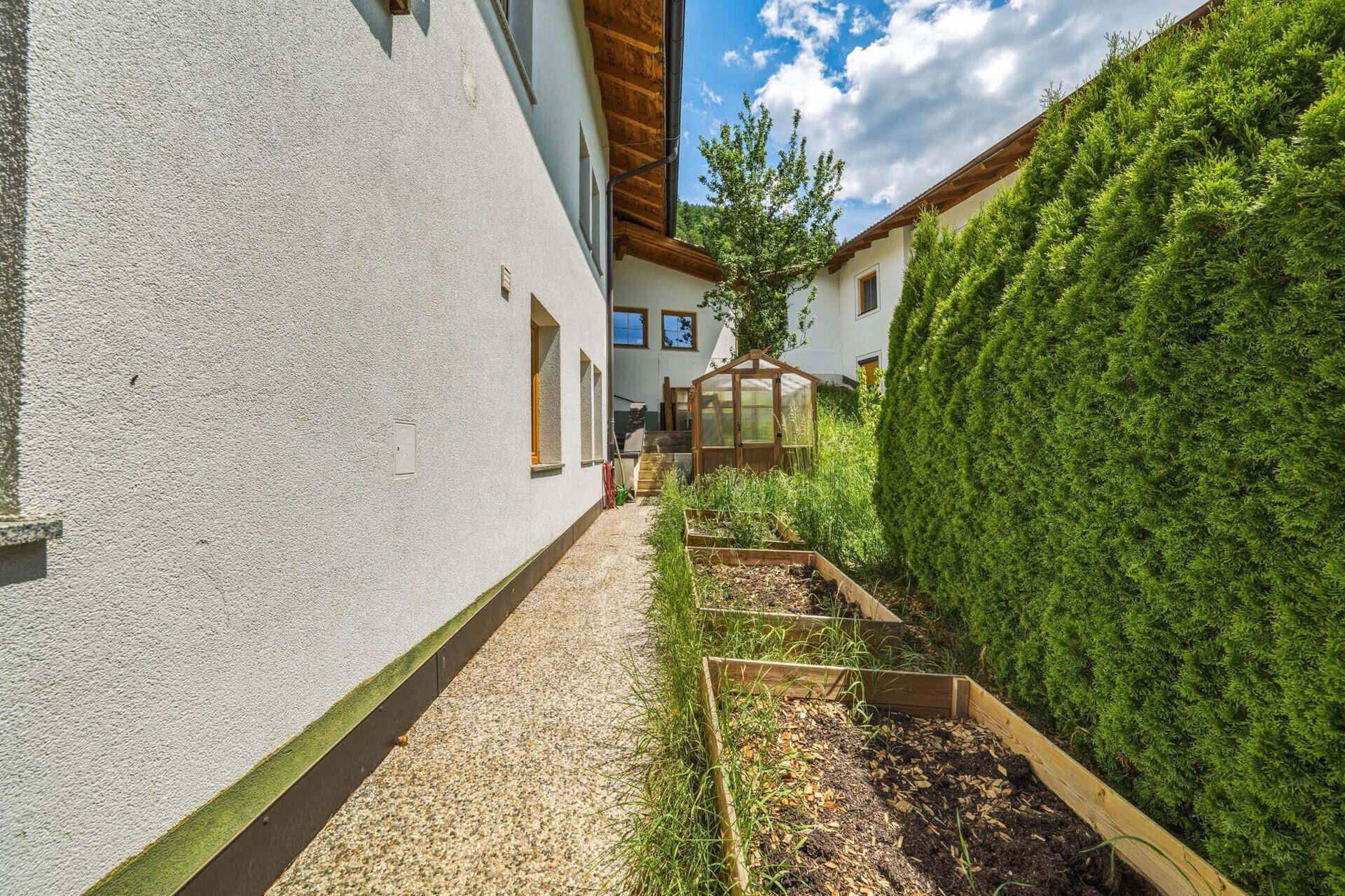 Schmaler Gartenweg entlang des Hauses mit Hochbeeten und einer hohen Hecke für Privatsphäre.