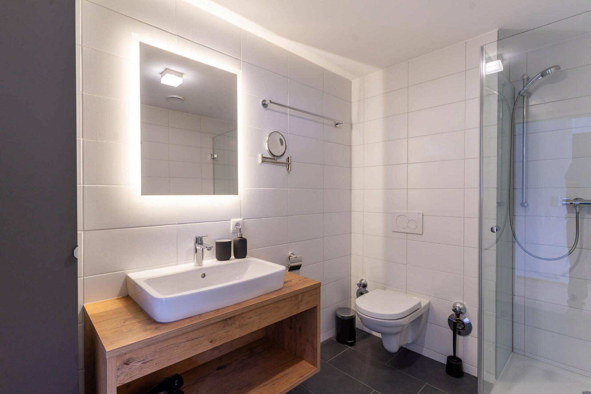 Modernes Badezimmer mit begehbarer Dusche, Holzwaschtisch und beleuchtetem Spiegel.