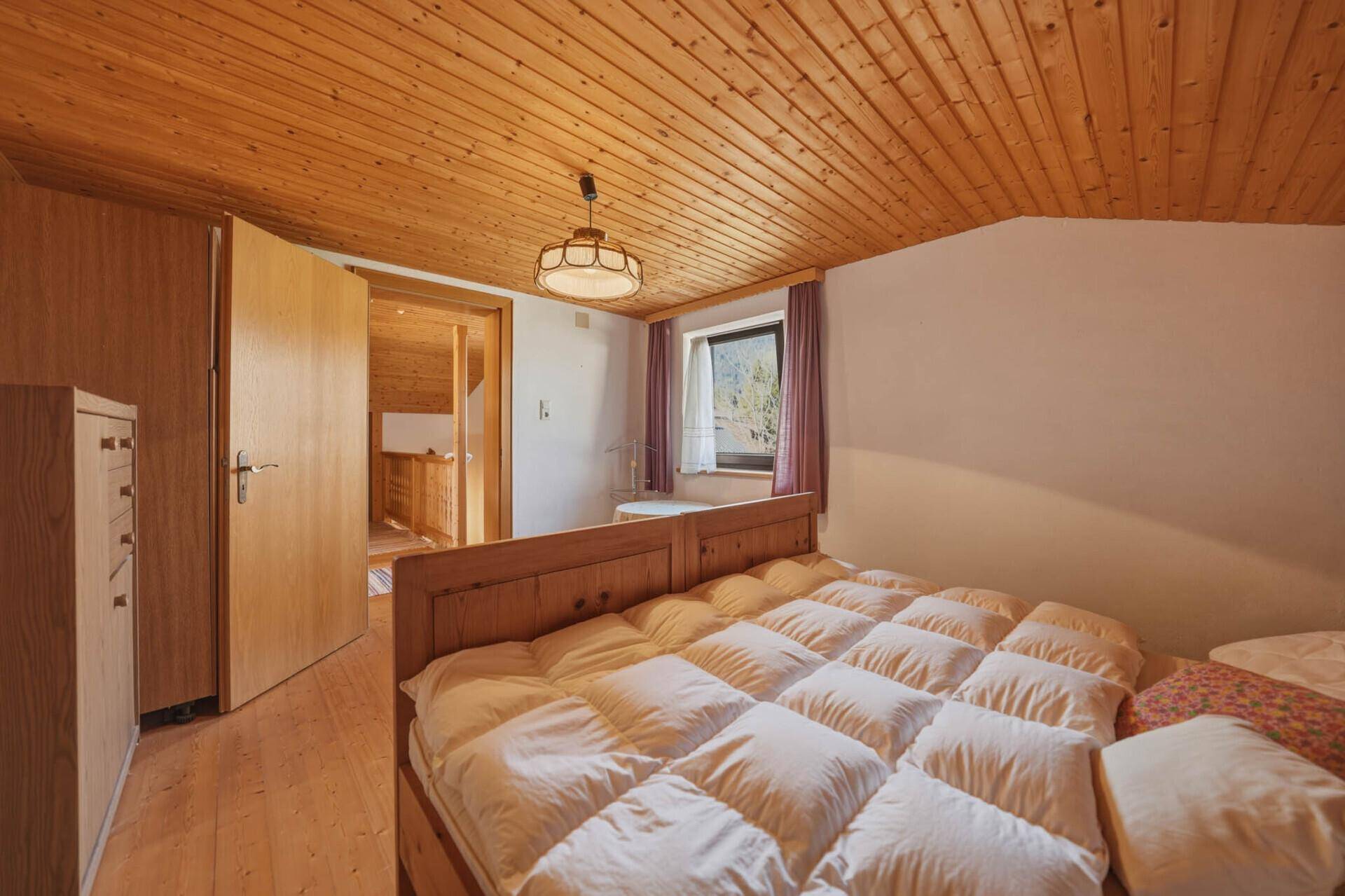 Helles Schlafzimmer im Dachgeschoss mit Holzdecke und -wänden, Doppelbett und Zugang zum Bad.