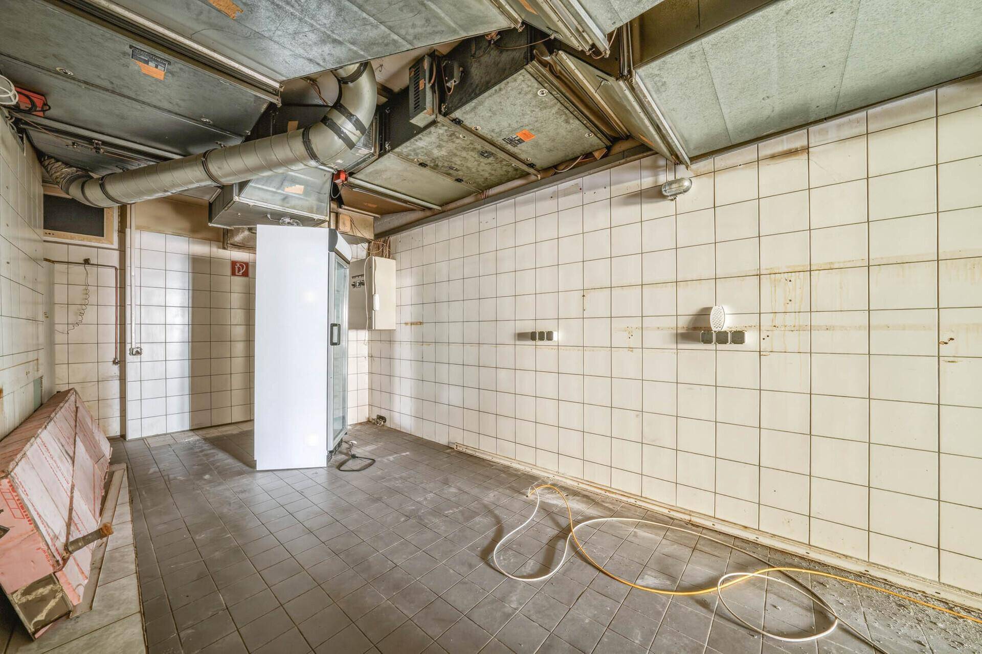 Industrielle Küche mit weißen Fliesen, sichtbaren Lüftungsrohren und einem Kühlschrank.