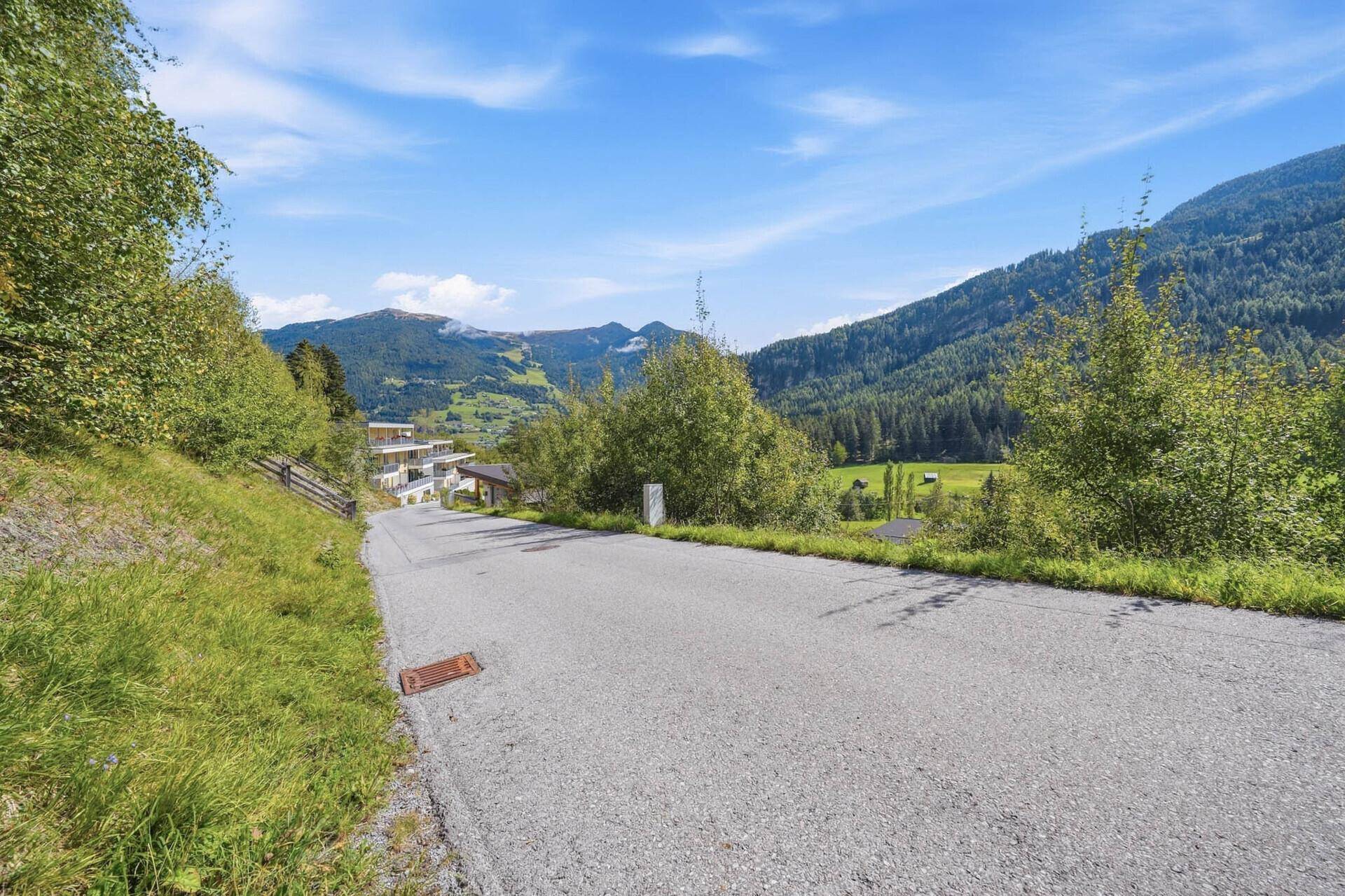 Eine ansteigende Straße führt durch eine grüne Landschaft mit Häusern und Bergen im Hintergrund.
