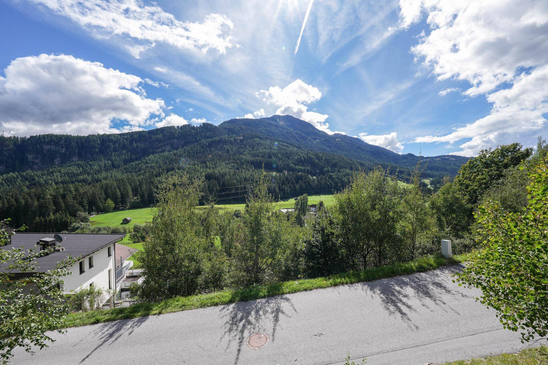 Blick von einer Landstraße auf bewaldete Berge und grüne Wiesen unter einem strahlend blauen Himmel.