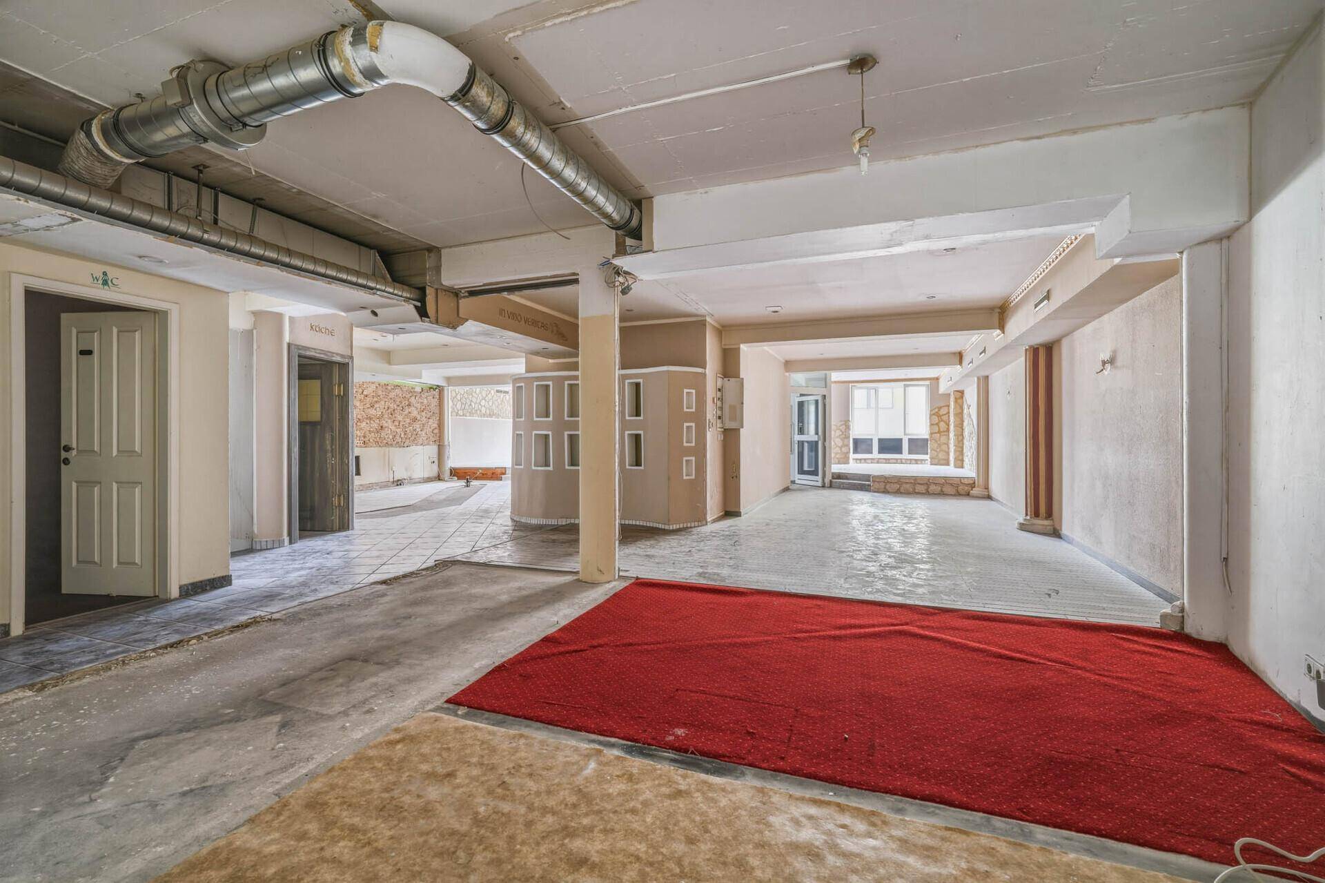 Großer Gewerberaum mit rotem Teppich, sichtbaren Lüftungsrohren und Zugang zu verschiedenen Bereichen.