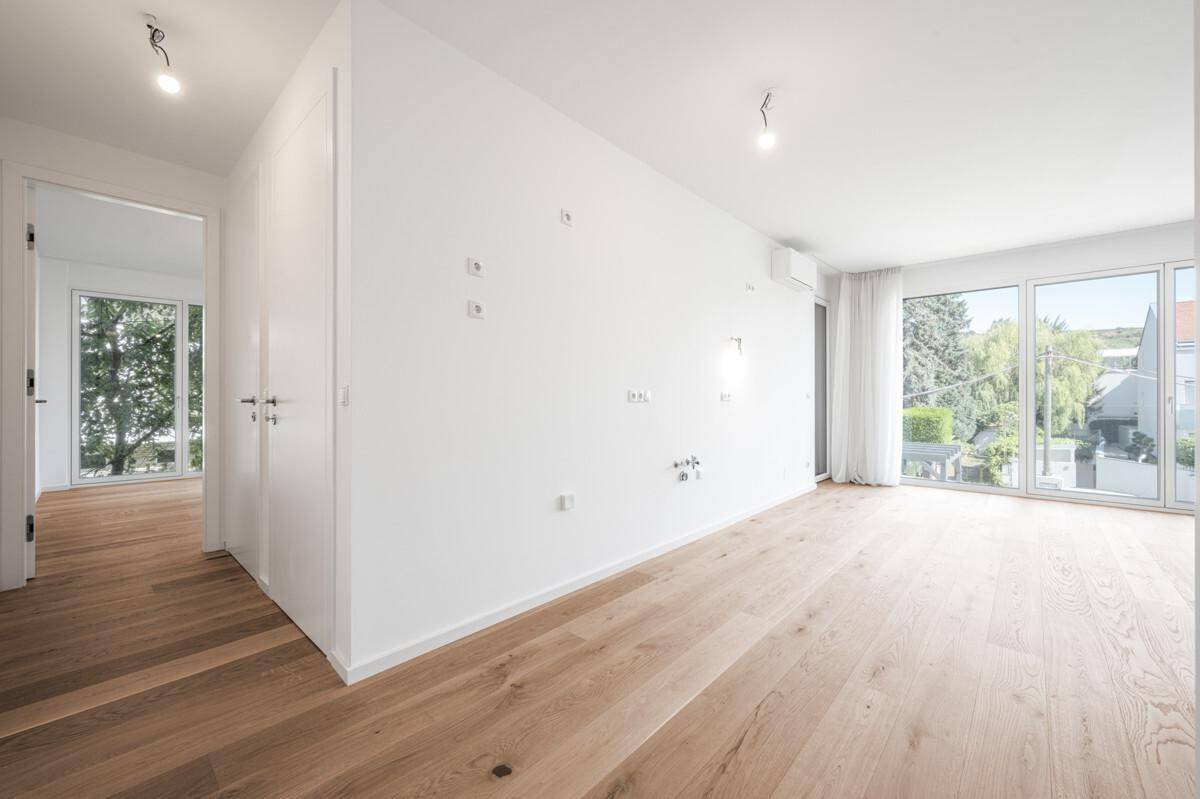 Heller Raum mit Holzboden, großen Fenstern und einem offenen Übergang zu weiteren Bereichen.