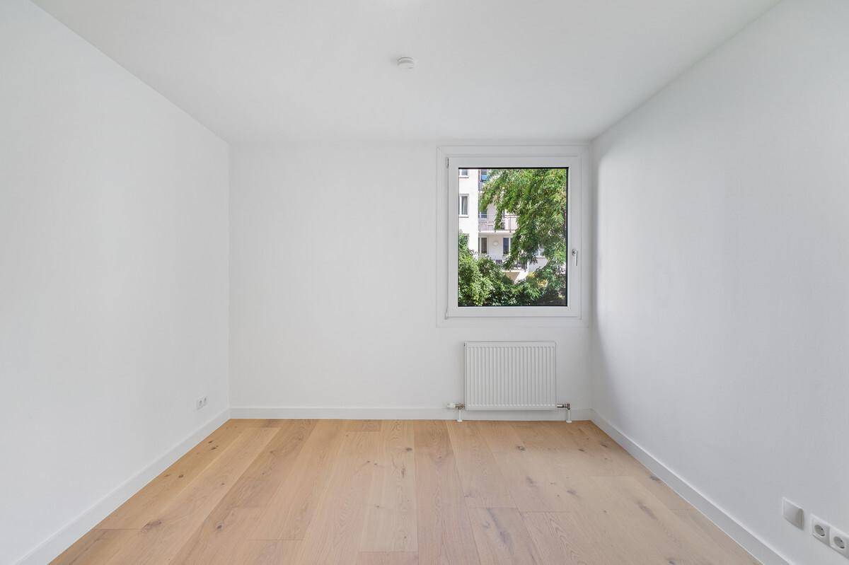 Helles Schlafzimmer mit Parkettboden und Fenster mit Blick auf Bäume und Gebäude.