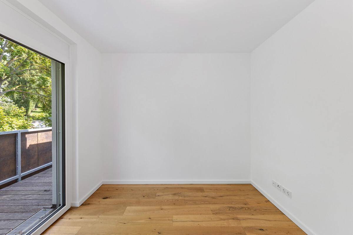 Leerer Raum mit Holzboden und Zugang zu einem Balkon, umgeben von Bäumen.