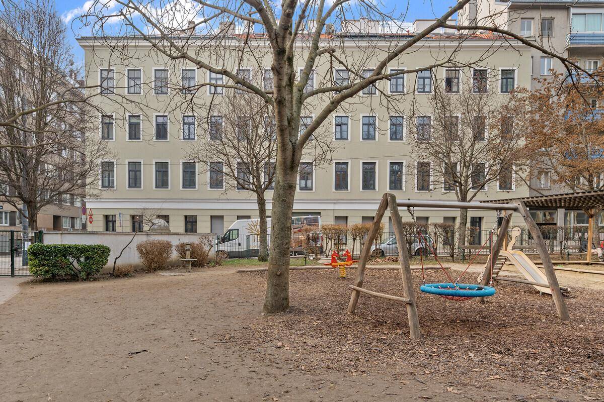 Rückansicht des Gebäudes mit einem angrenzenden Spielplatz und Bäumen im Vordergrund.