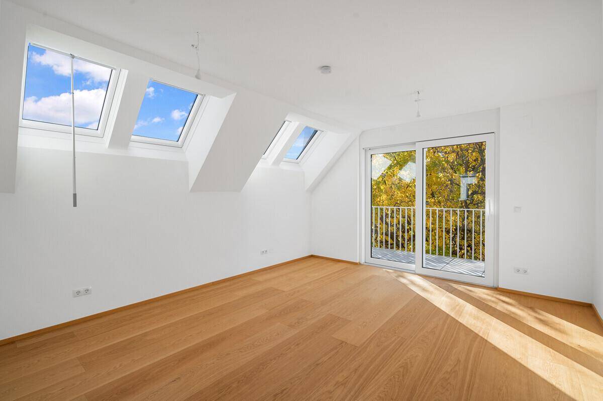 Heller Raum mit Holzboden, Dachschrägen, mehreren Dachfenstern und Balkonzugang.