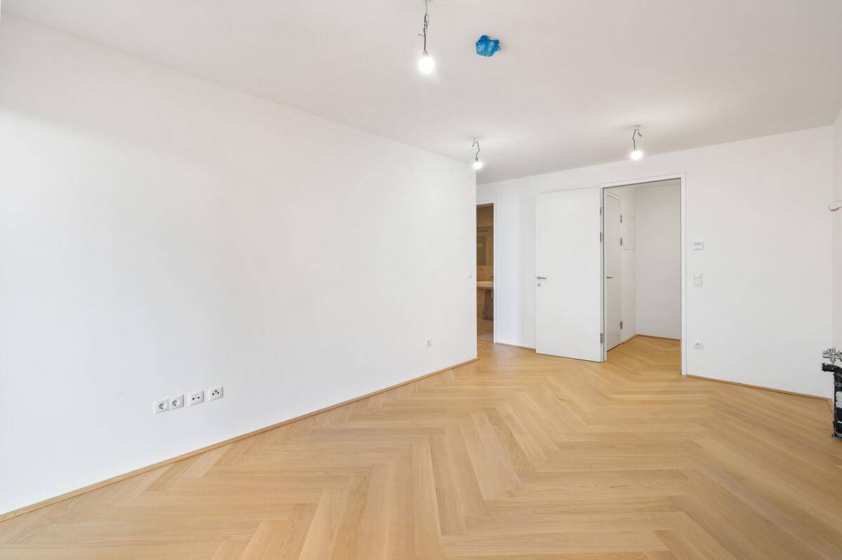 Geräumiger, unmöblierter Raum mit hellem Fischgrätparkett und offener Tür.