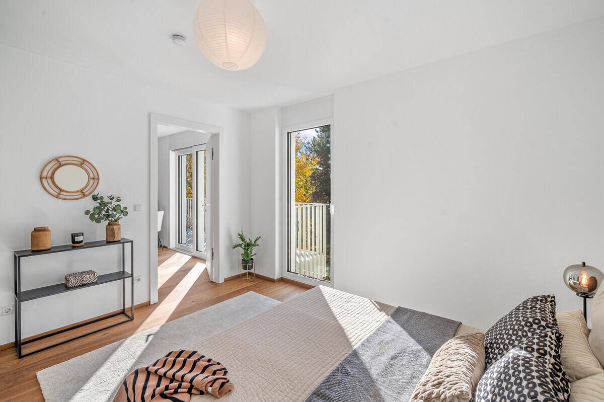 Heller Raum mit Holzboden, einem Fenster und Zugang zu einem Balkon mit grüner Aussicht.