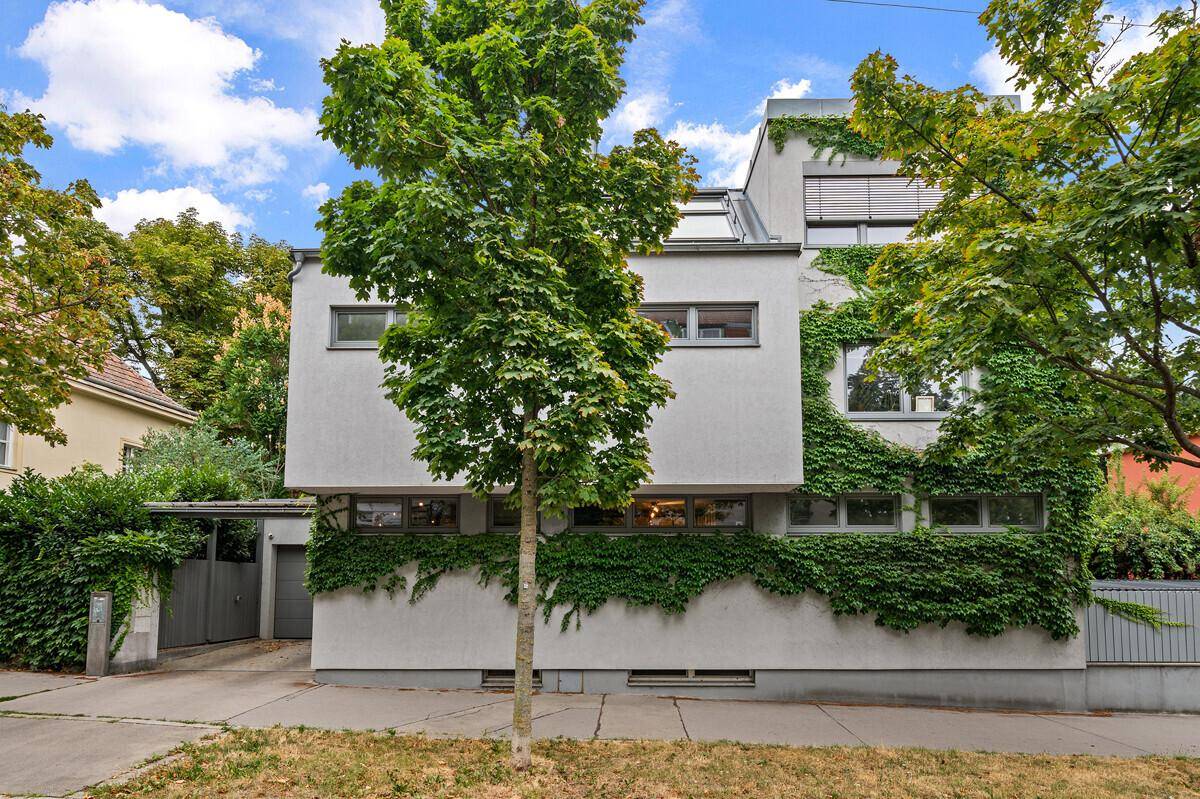 Ansicht der modernen Hausfassade mit üppiger Begrünung und großen Fenstern, umgeben von Bäumen.