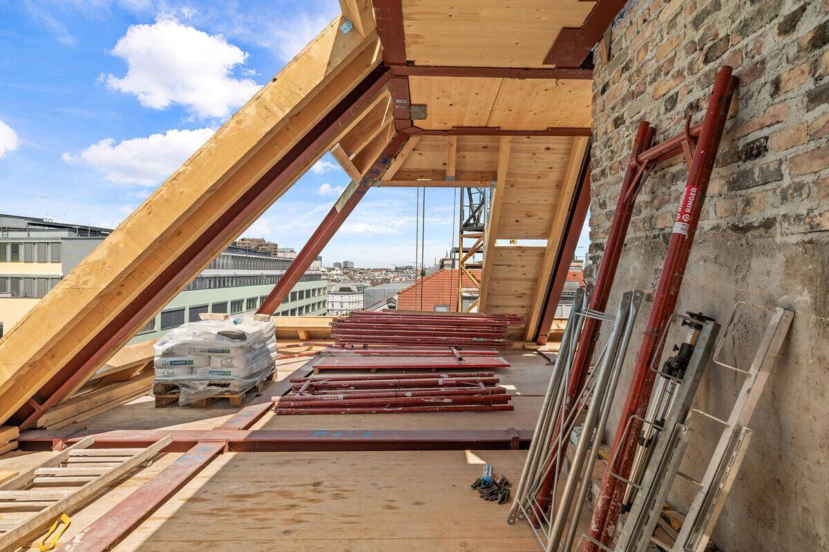Dachgeschoss im Rohbau mit sichtbaren Holzbalken und Baumaterialien unter blauem Himmel.