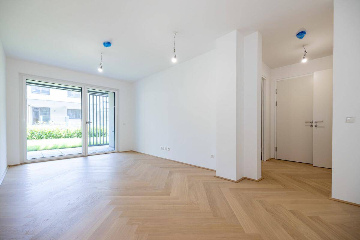 Heller, leerer Raum mit Fischgrätparkett und großer Fenstertür zum Außenbereich.