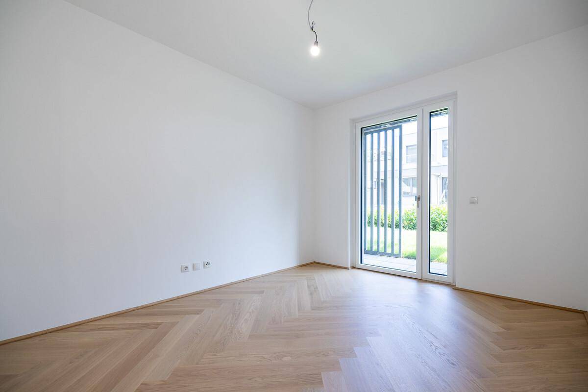 Leeres Zimmer mit hellem Fischgrätparkett und großer Fenstertür zum Außenbereich.