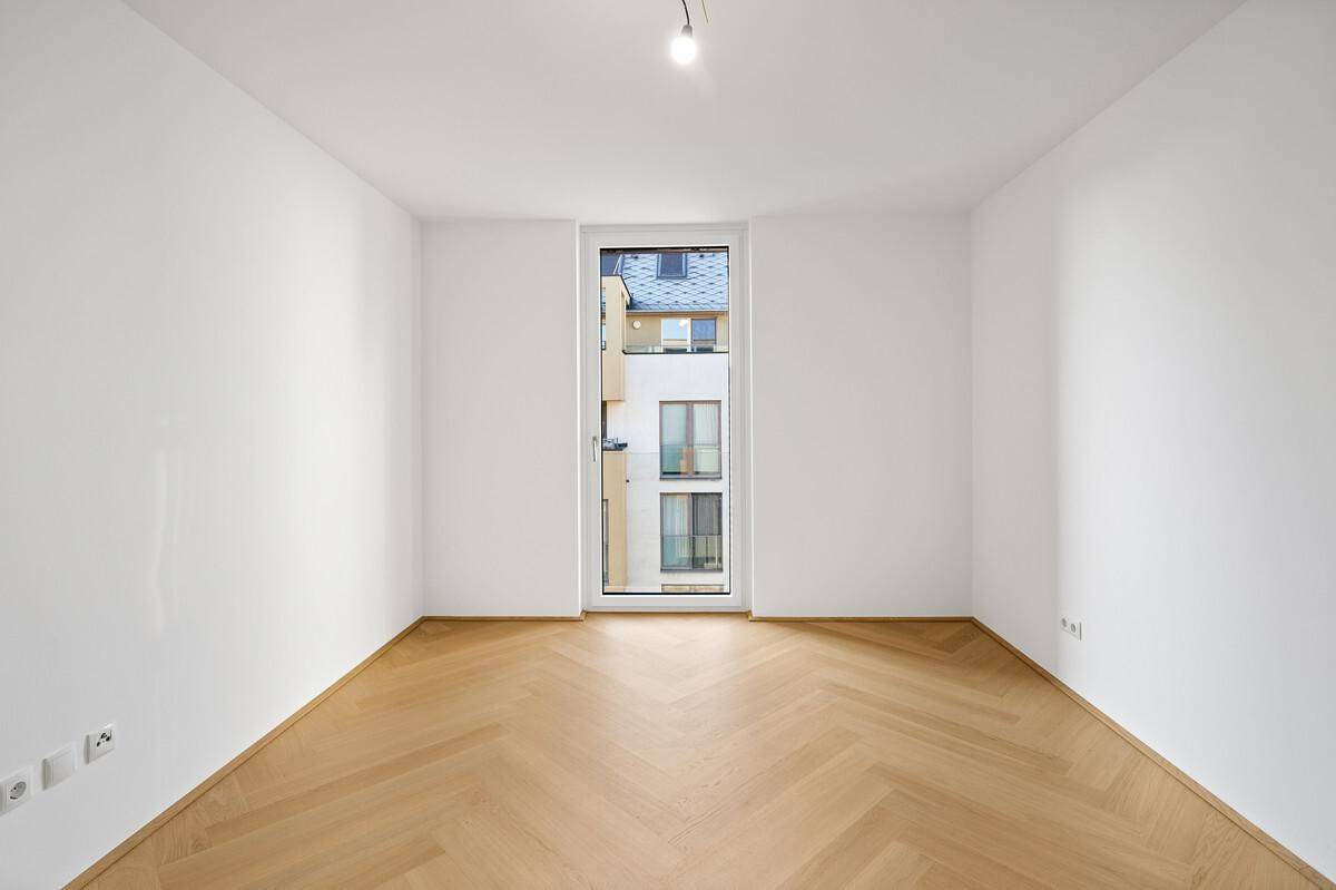 Leeres Schlafzimmer mit Fischgrätparkett und einem hohen Fenster mit Blick nach draußen.