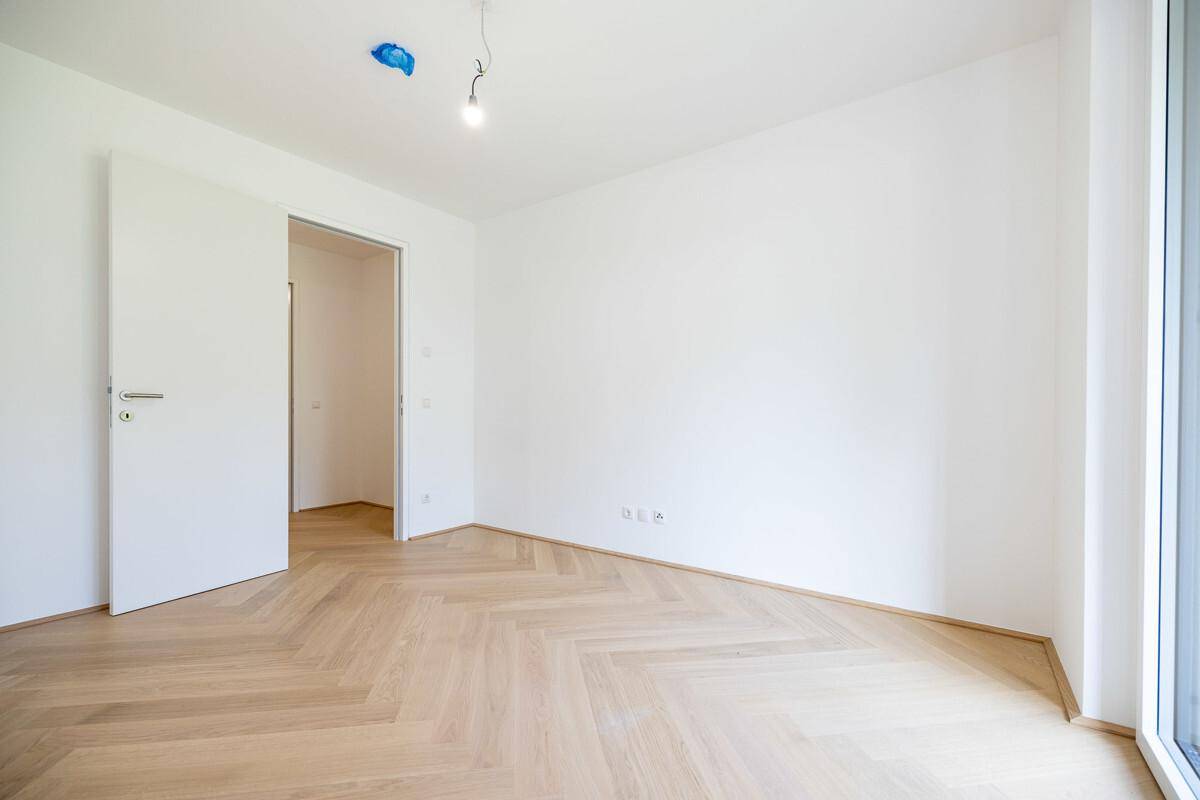 Unmöbliertes Zimmer mit hellem Fischgrätparkett und weiß gestrichenen Wänden.
