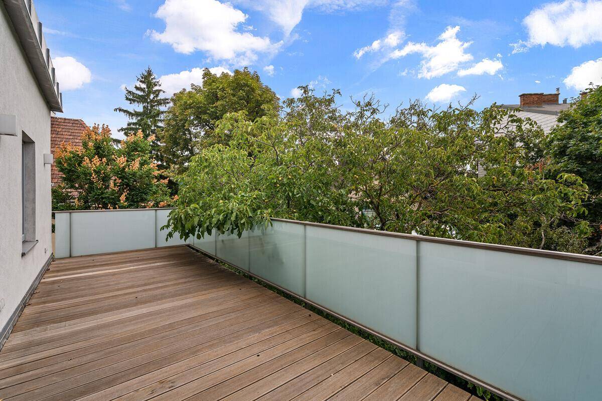 Geräumiger Balkon mit Holzboden und Milchglasgeländer, umgeben von Bäumen und viel Grün.
