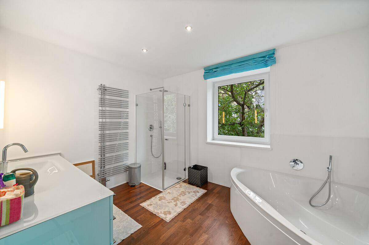 Helles Badezimmer mit Badewanne, Duschkabine, beheiztem Handtuchhalter und Fenster mit Grünblick.