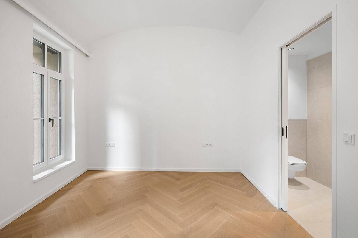 Leeres Zimmer mit Fischgrätparkett und einem Fenster, das direkt zu einem modernen Bad führt.