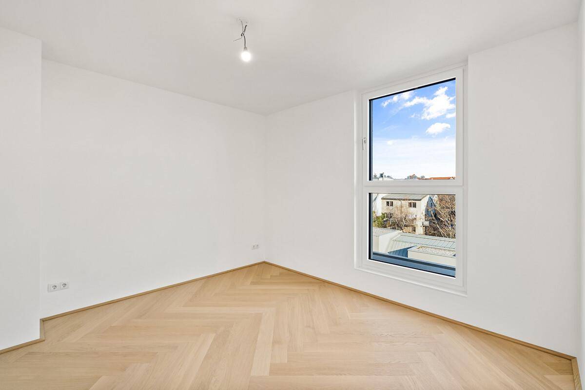 Heller Raum mit Fischgrätparkett und einem großen Fenster, das viel Tageslicht hereinlässt.