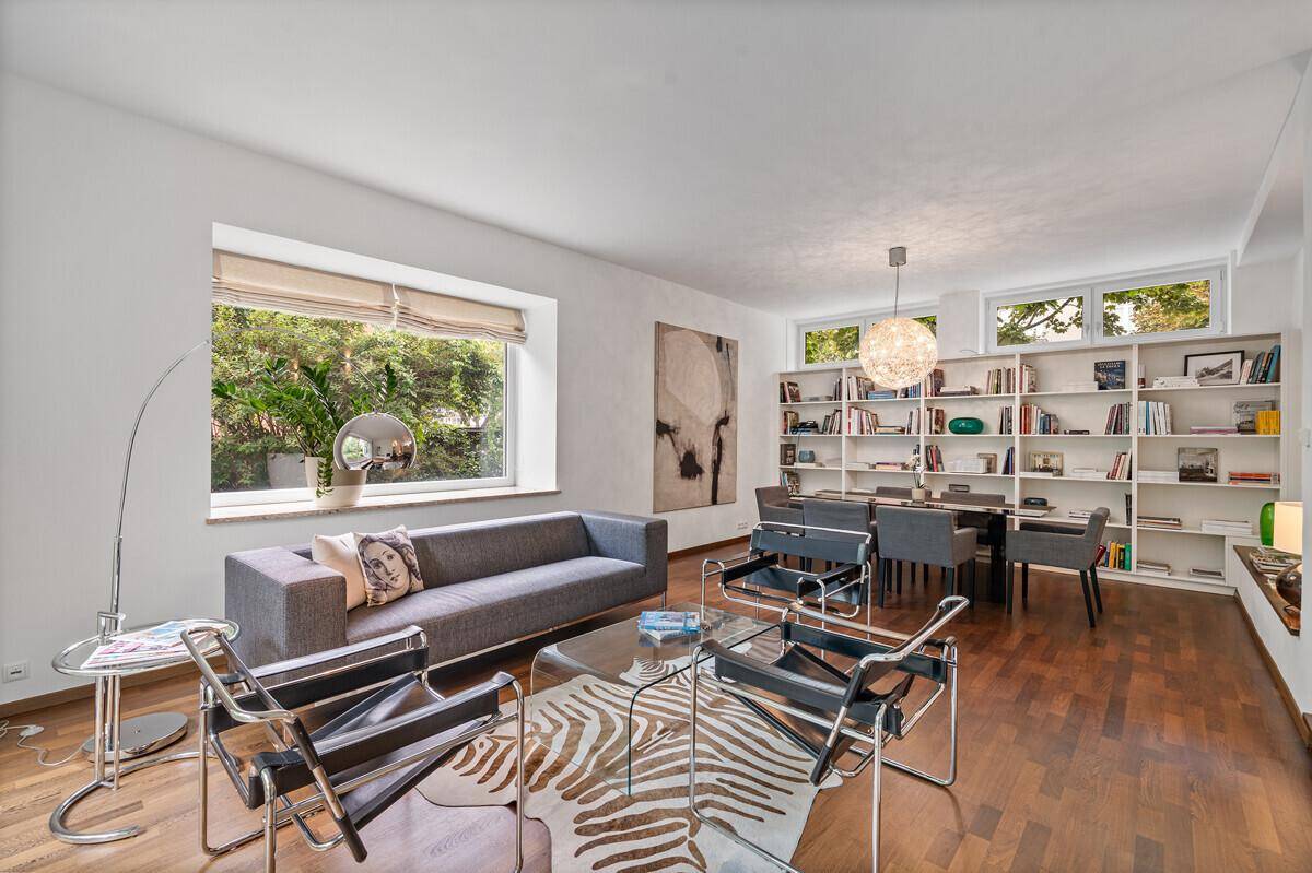 Geräumiger Wohn- und Essbereich mit Bücherregalen, Holzboden und viel Tageslicht durch große Fenster.