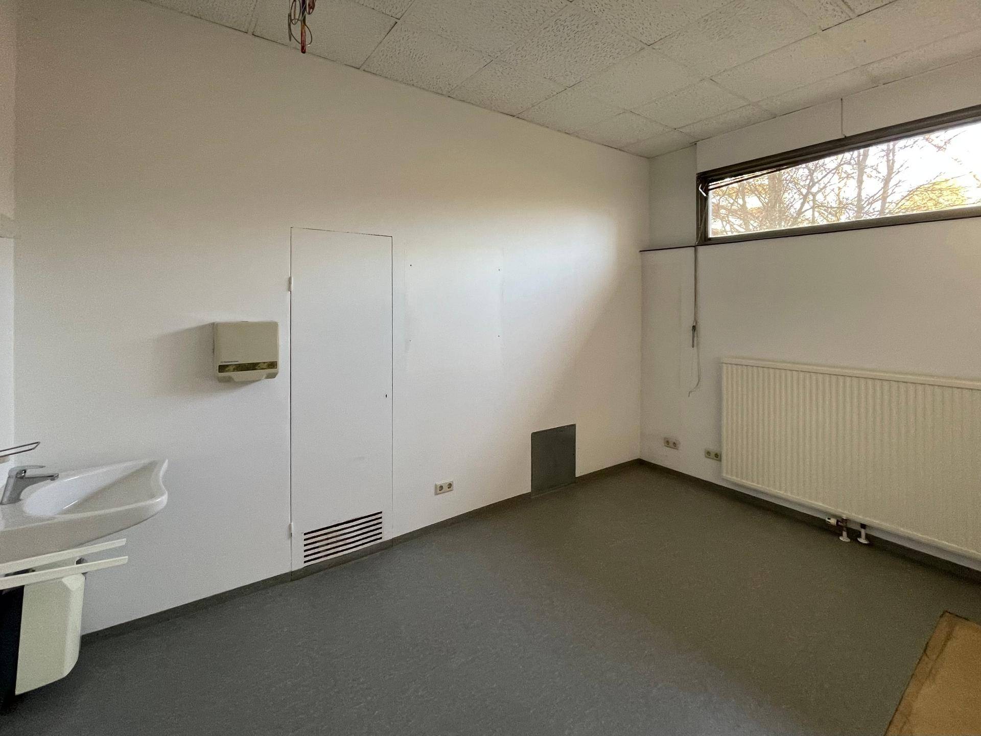 Kleiner Büroraum mit Waschbecken, Heizkörper und einem hohen Fenster für Tageslicht.