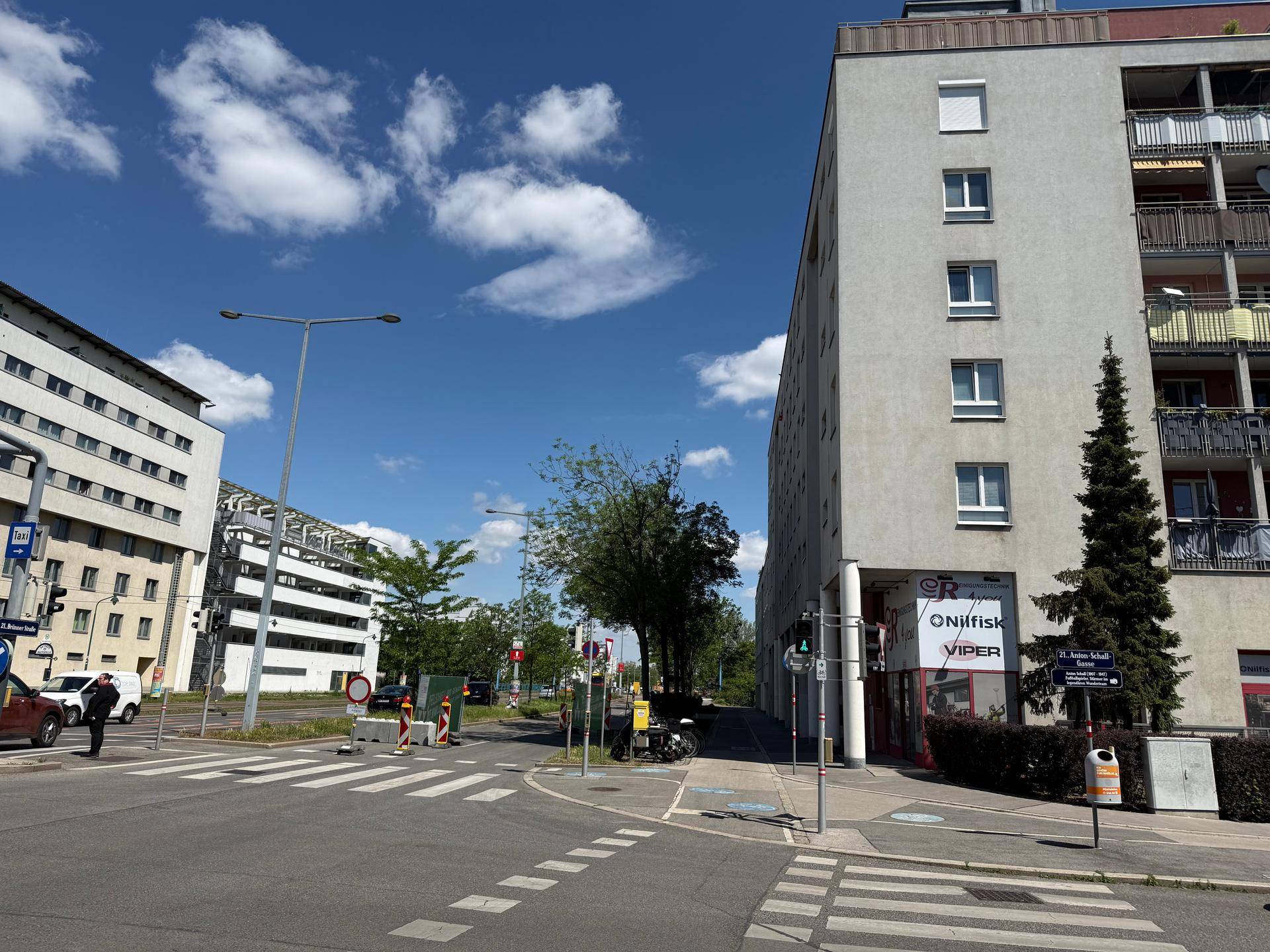 Weitläufige Straßenansicht mit einem Mehrfamilienhaus, Geschäften im Erdgeschoss und viel Grün.