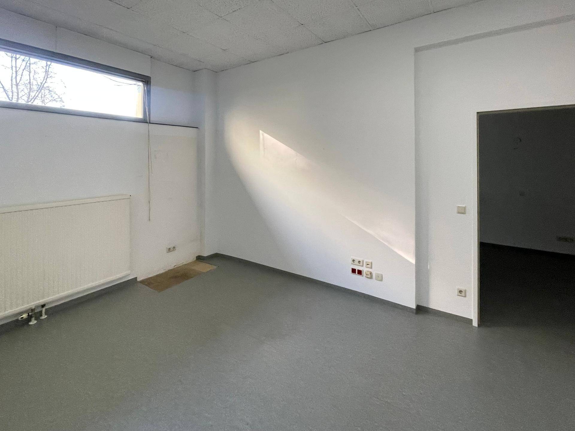 Leerer Büroraum mit einem hohen Fenster, Heizkörper und mehreren sichtbaren Steckdosen.