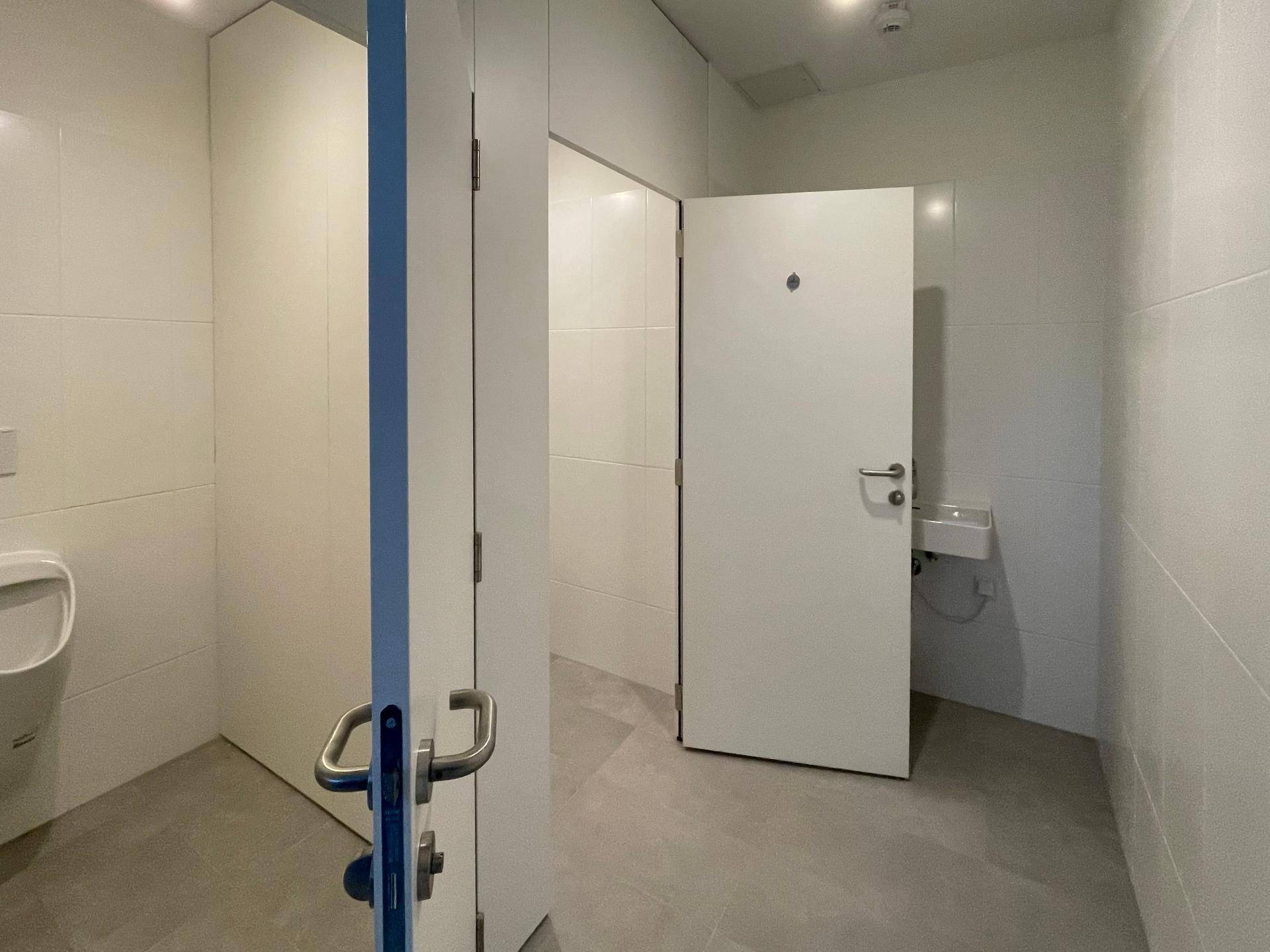 Modernes und helles öffentliches Badezimmer mit mehreren Kabinen, Urinal und weißen Fliesen.