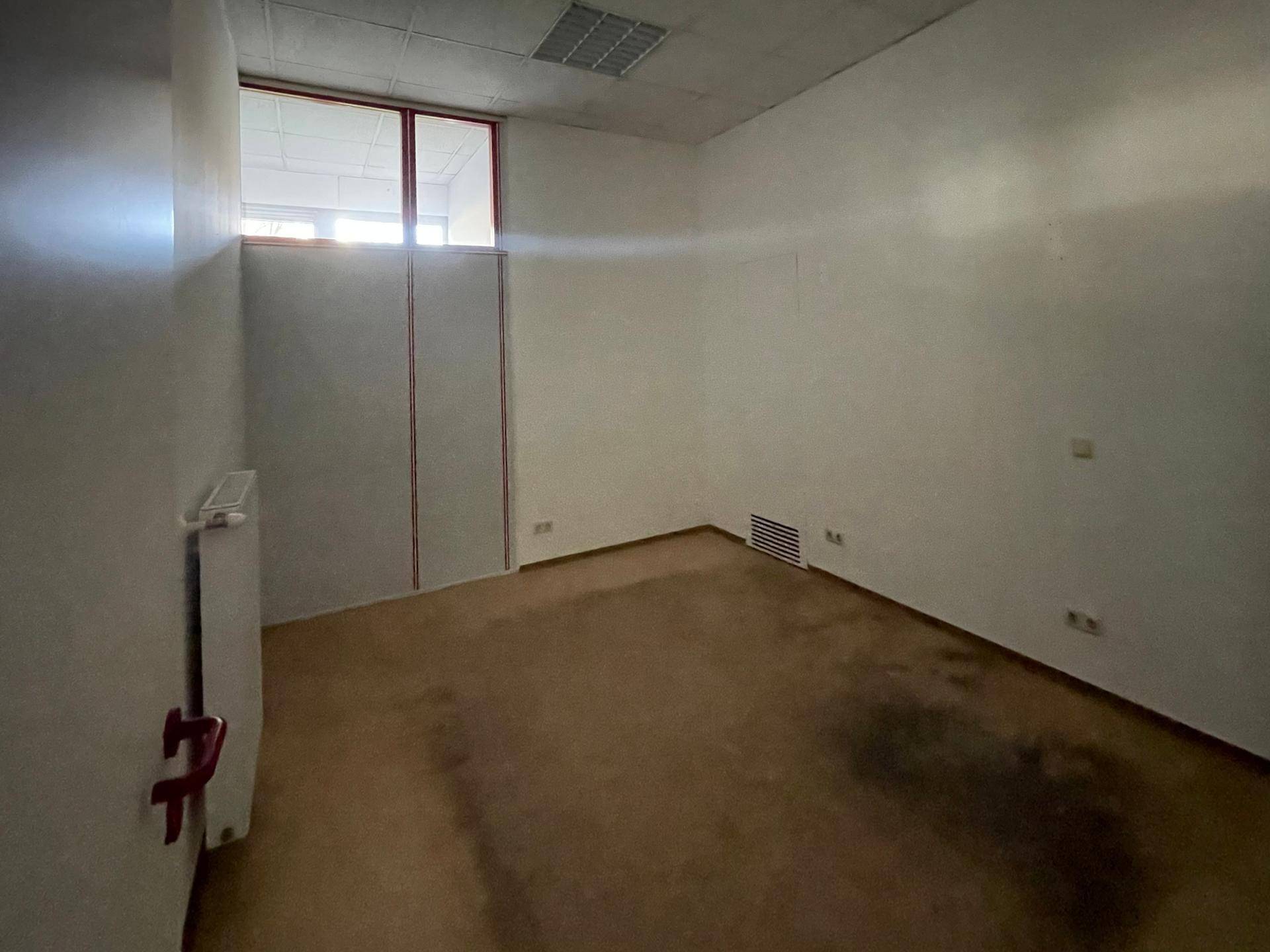 Leerer Büroraum mit einem hohen Fenster, Heizkörper und einem braunen Bodenbelag.