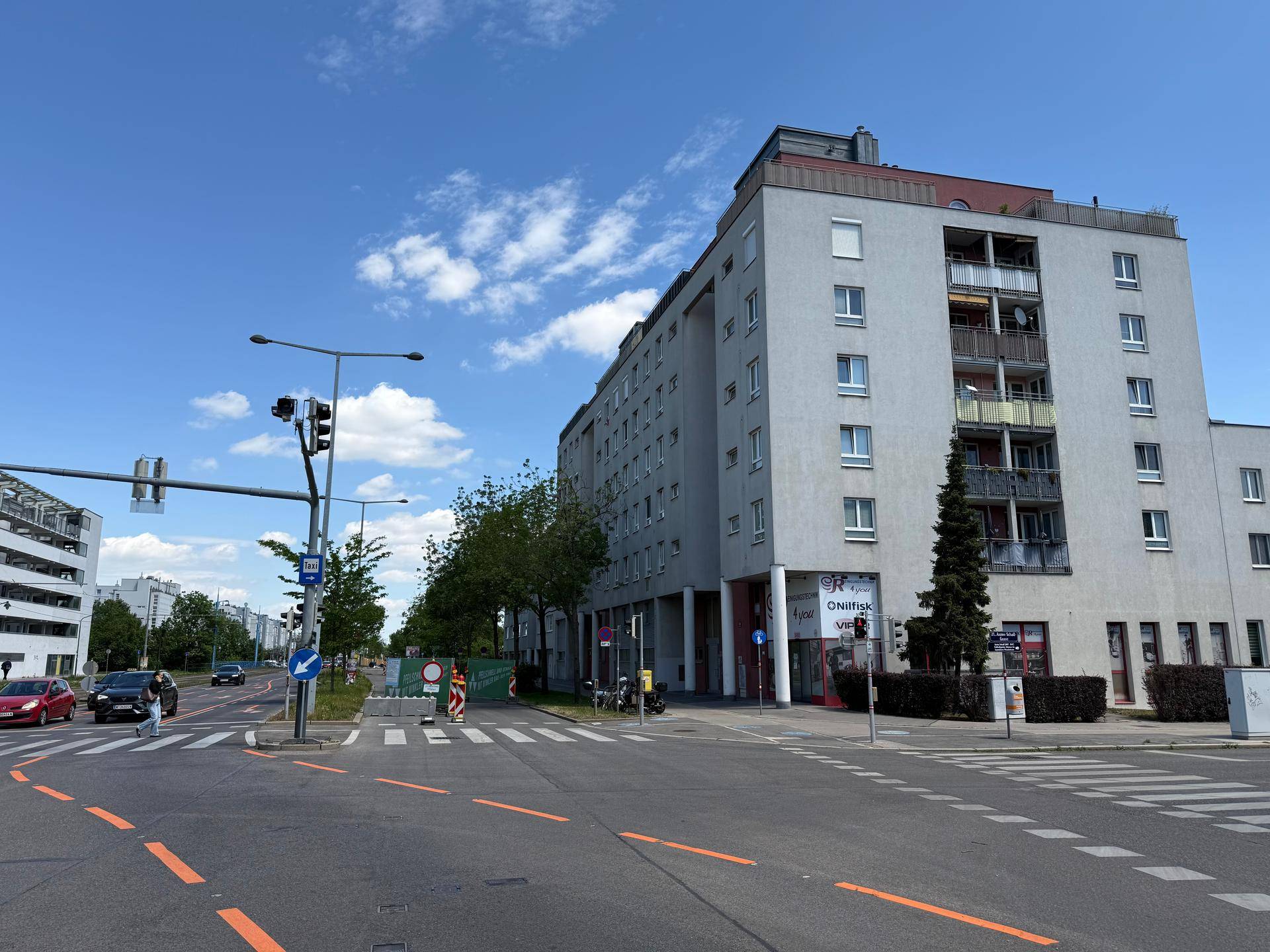 Panoramablick auf eine belebte Straße mit einem großen Wohn- und Geschäftsgebäude unter blauem Himmel.