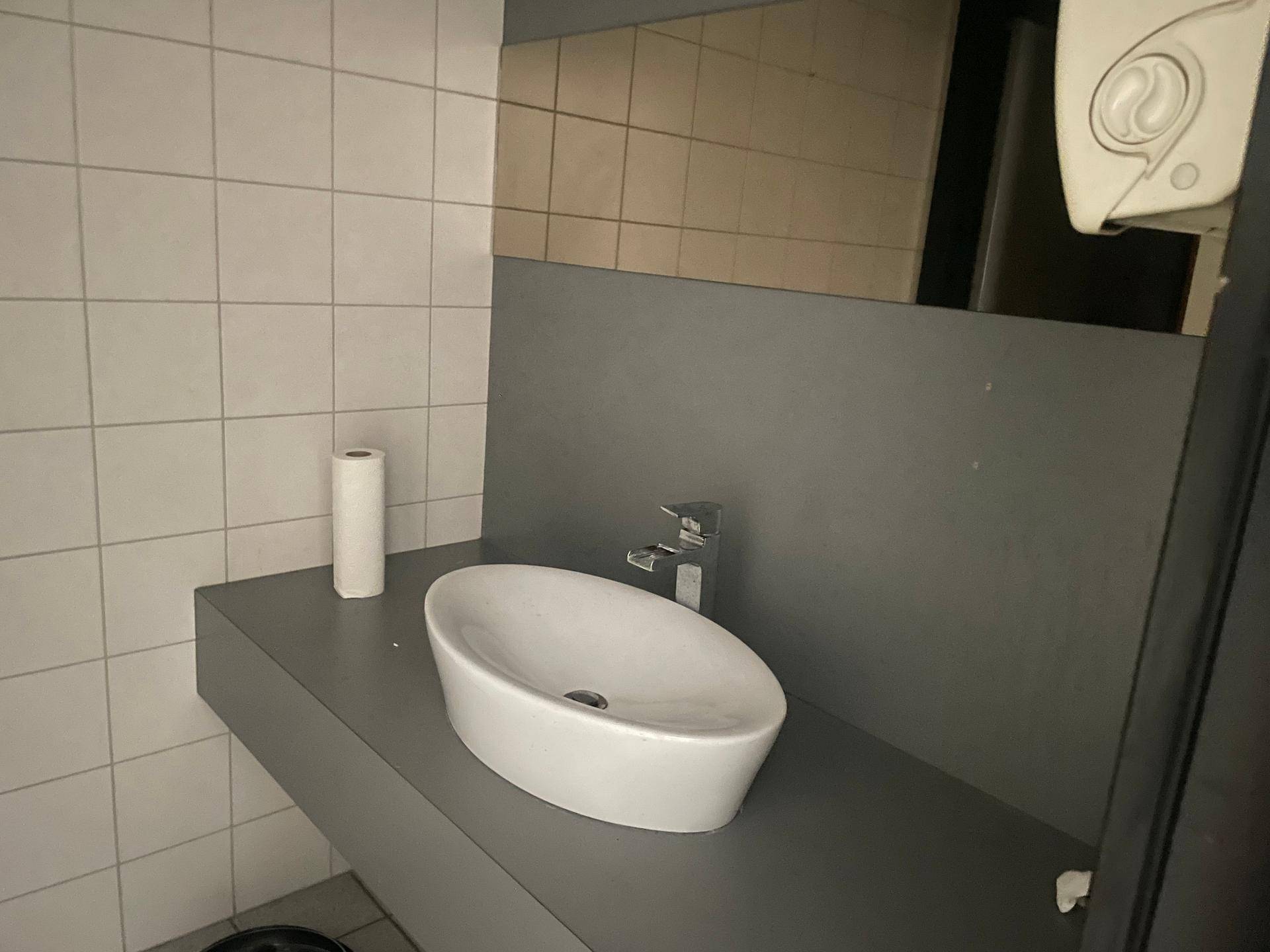 Modernes Waschbecken auf einer grauen Ablagefläche in einem gefliesten Toilettenraum mit Spiegel.