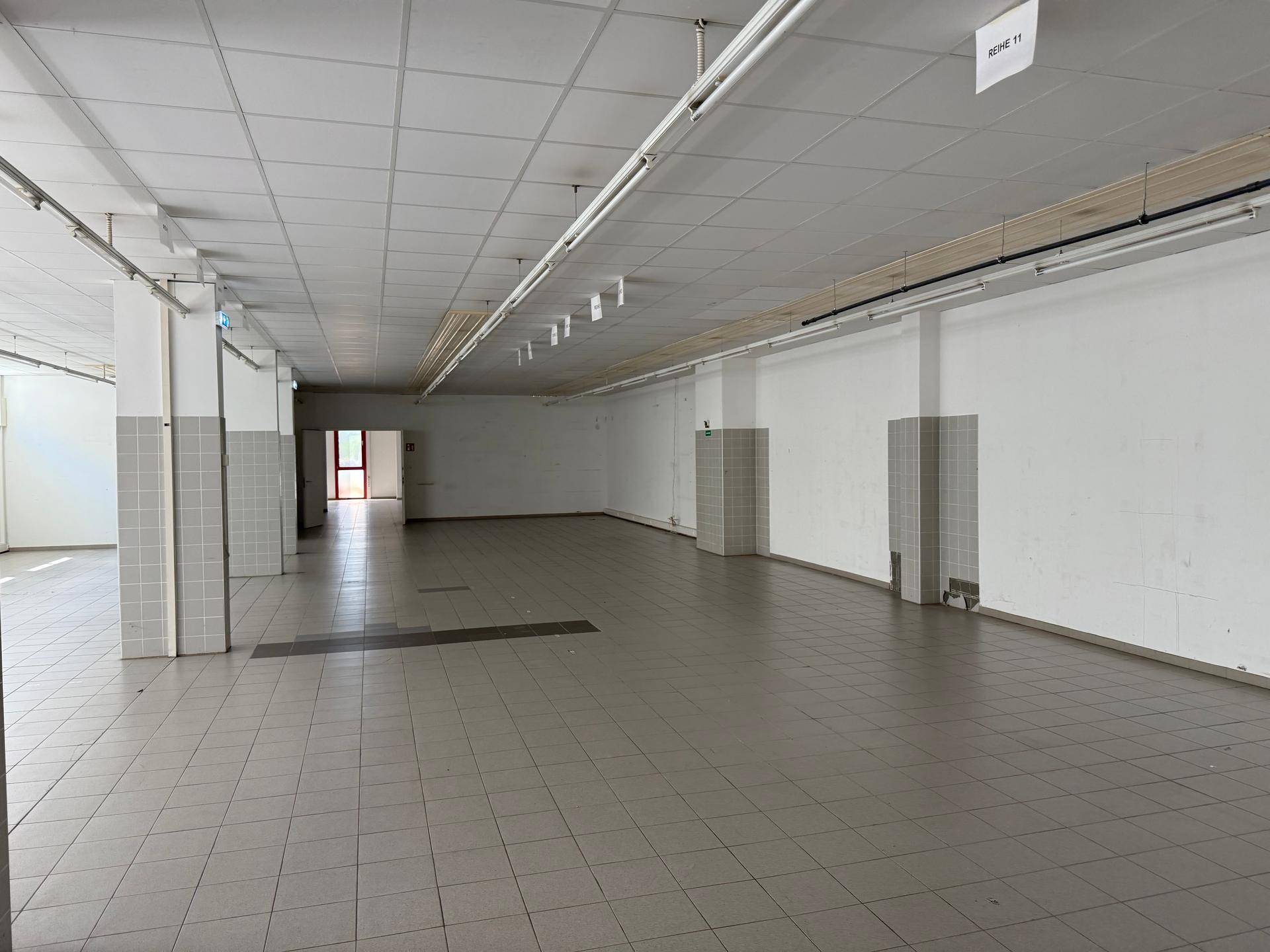 Weitläufiger, leerer Gewerberaum mit Fliesenboden, Säulen und Deckenbeleuchtung, bereit zur Gestaltung.