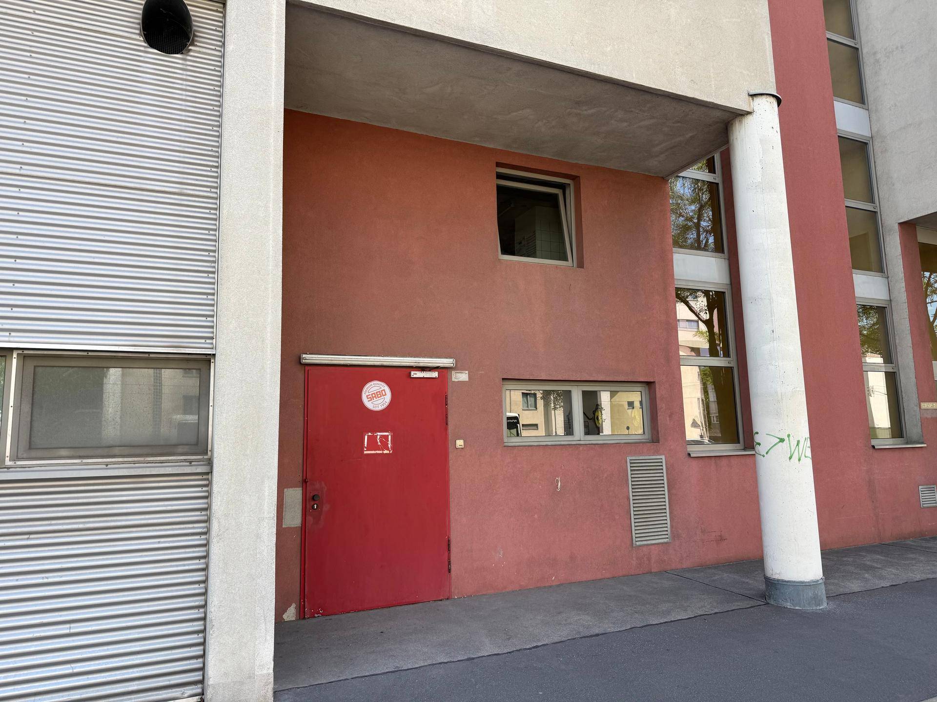 Roter Eingangsbereich eines Gebäudes mit Rolltor und Fenstern in einer modernen Fassade.