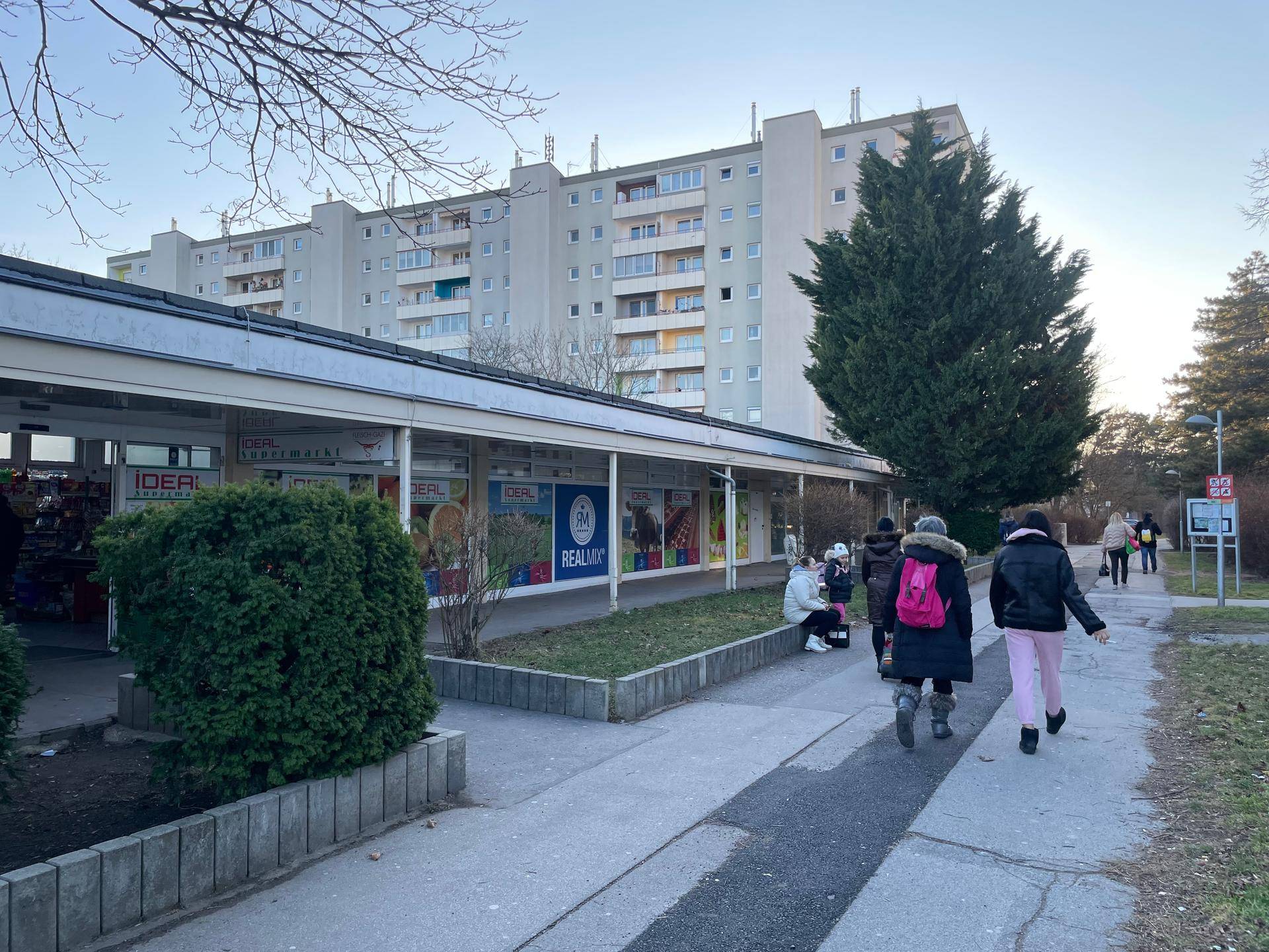 Blick auf ein großes Wohngebäude mit Geschäften im Erdgeschoss und einem belebten Gehweg davor, umgeben von Bäumen.